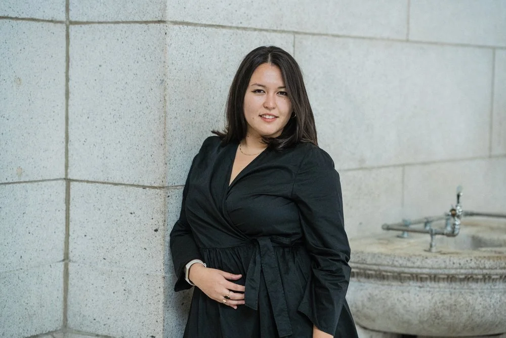 A woman with shoulder-length dark hair standing next to a concrete wall and a water fountain, wearing a black dress and smiling.