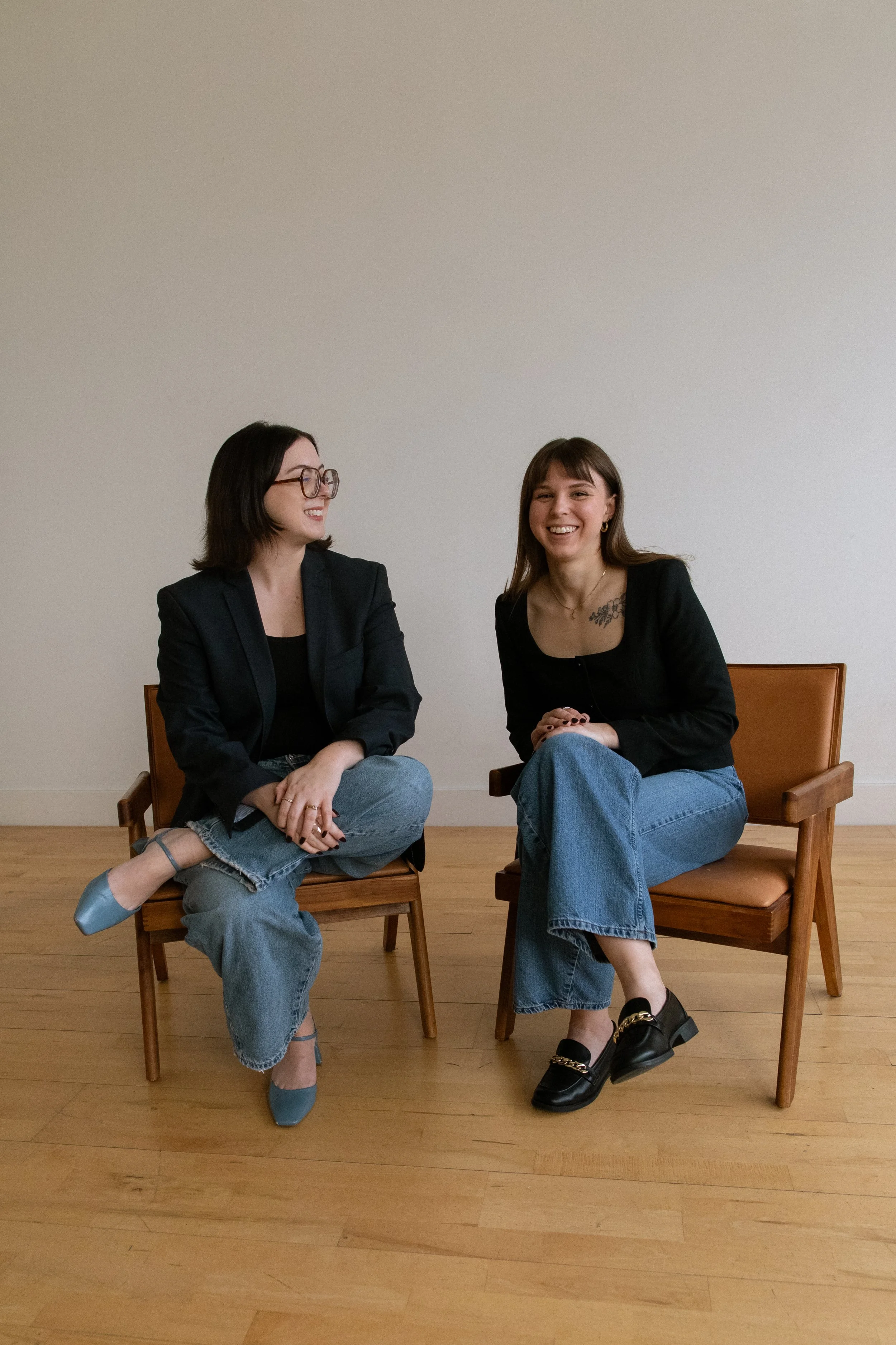Two women sitting on wooden chairs, laughing and engaging in conversation against a plain white wall.