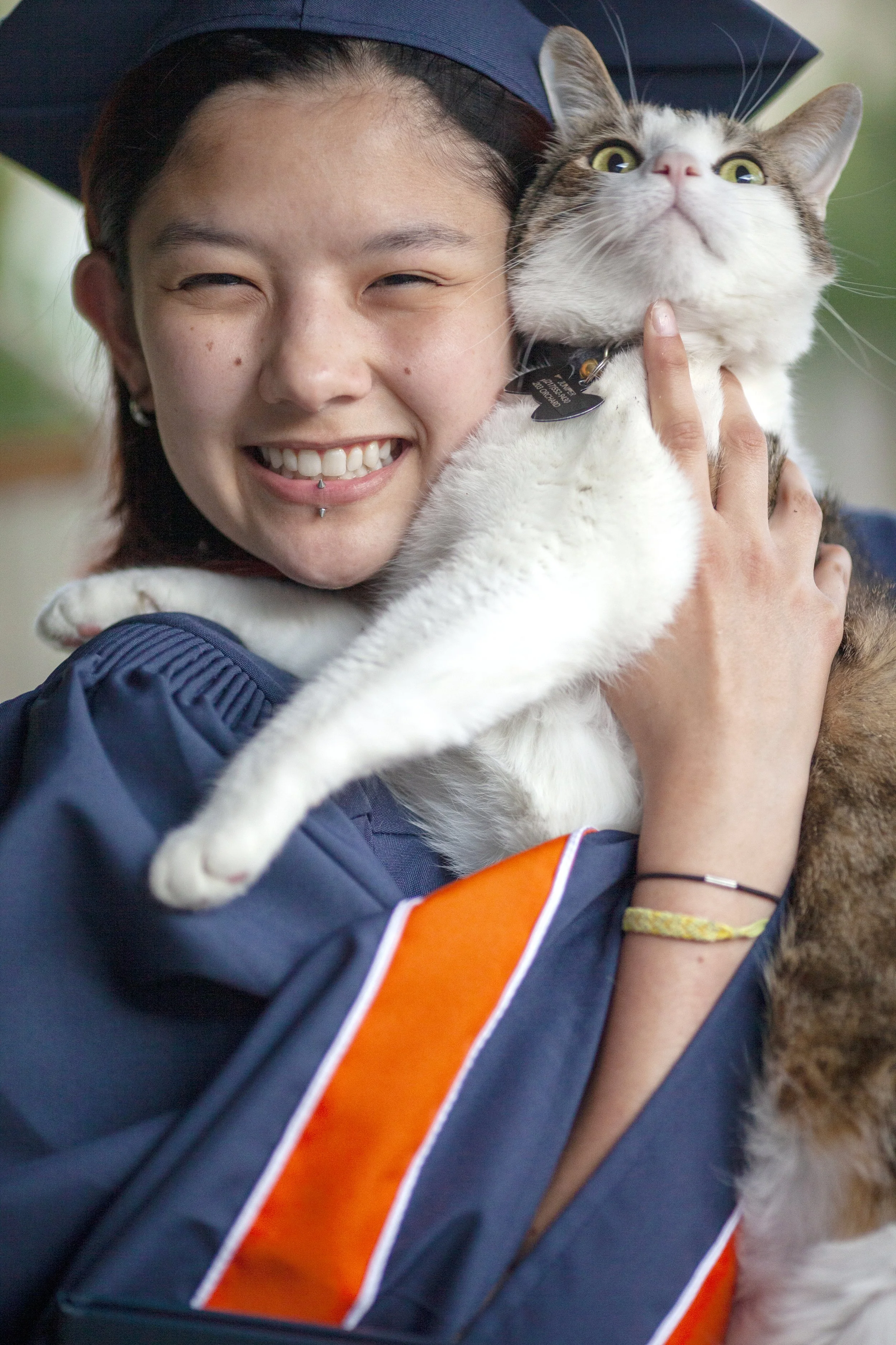 A young woman in a graduation cap and gown holding a large white and gray tabby cat, smiling happily.
