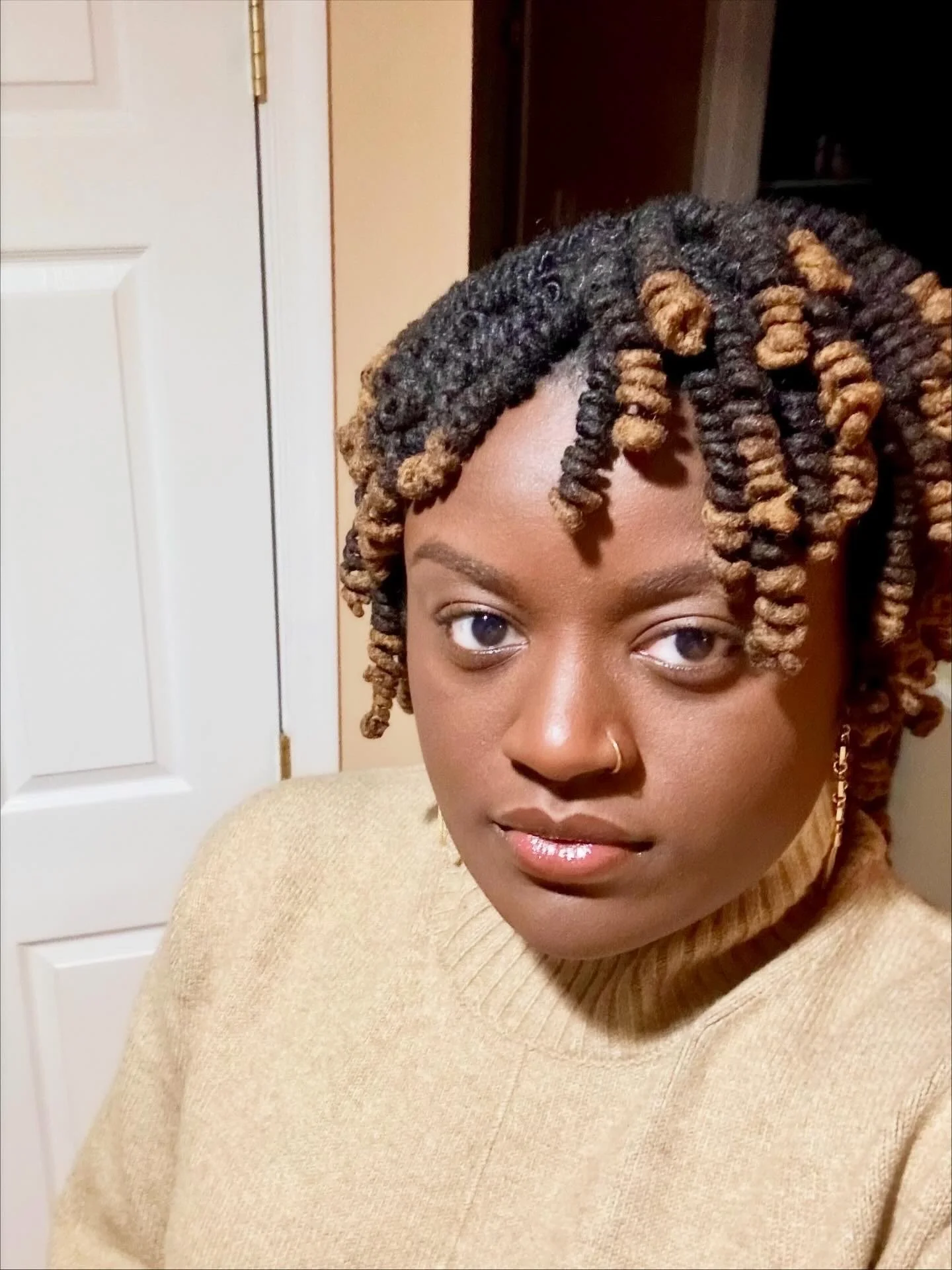 A young woman with styled dark and blonde twisted locks, wearing a beige sweater, a nose ring, and a long earring, standing indoors in front of a white door.