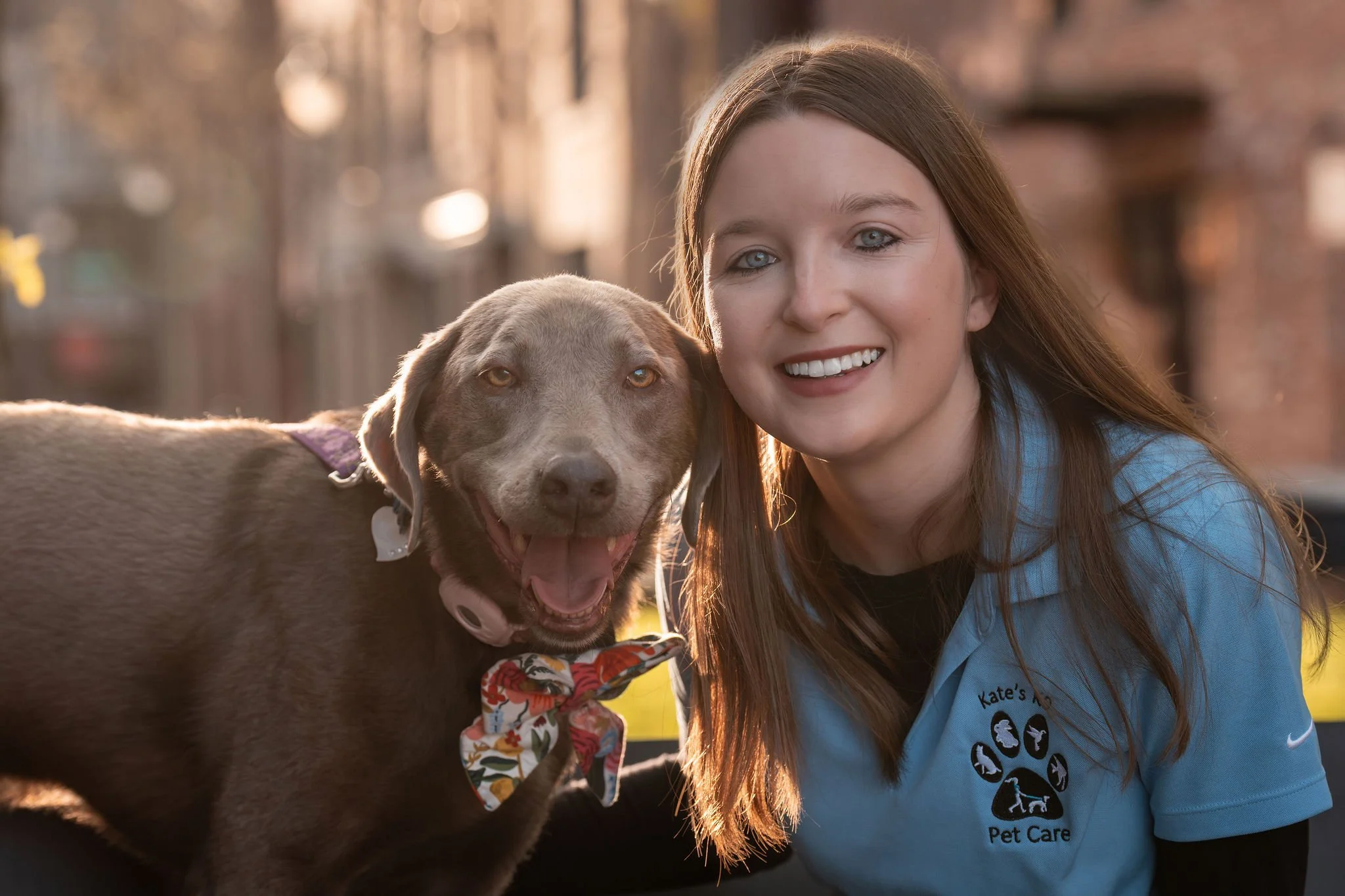 A woman with long brown hair and a blue pet care uniform smiling next to a happy brown dog wearing a floral bow tie, outdoors in a backyard with sunlight and trees in the background.
