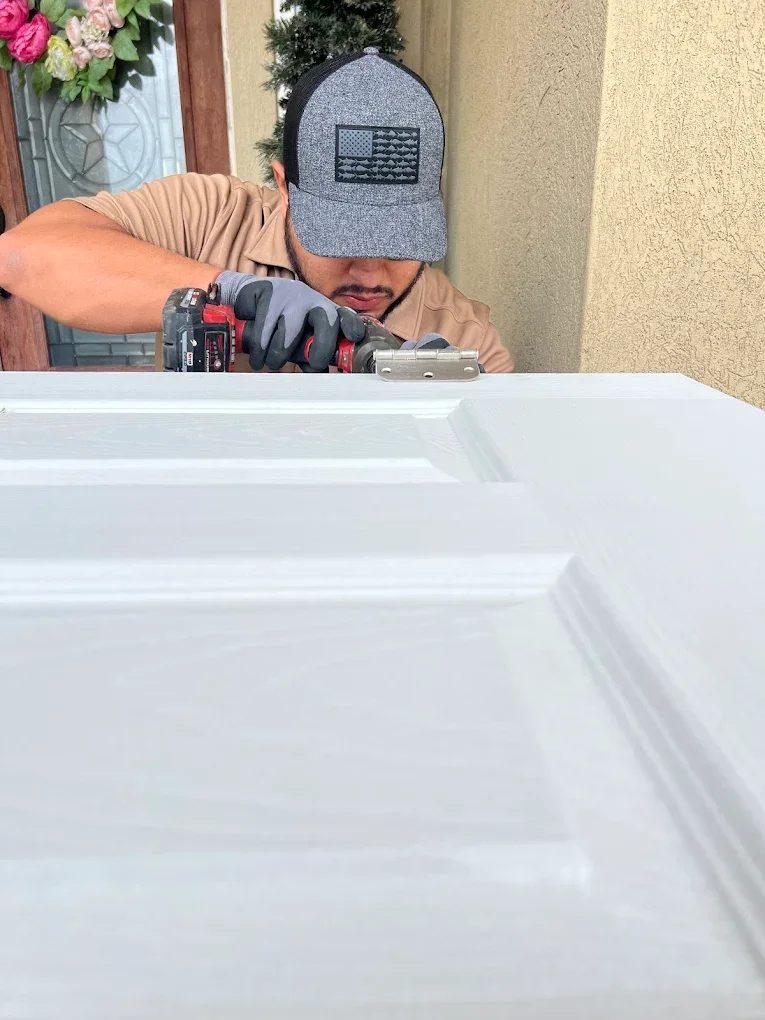 A man wearing a gray baseball cap with an American flag patch and a brown shirt is using a cordless drill to work on a white surface, likely a door, outside a house with a yellow wall and a flower arrangement in the background.