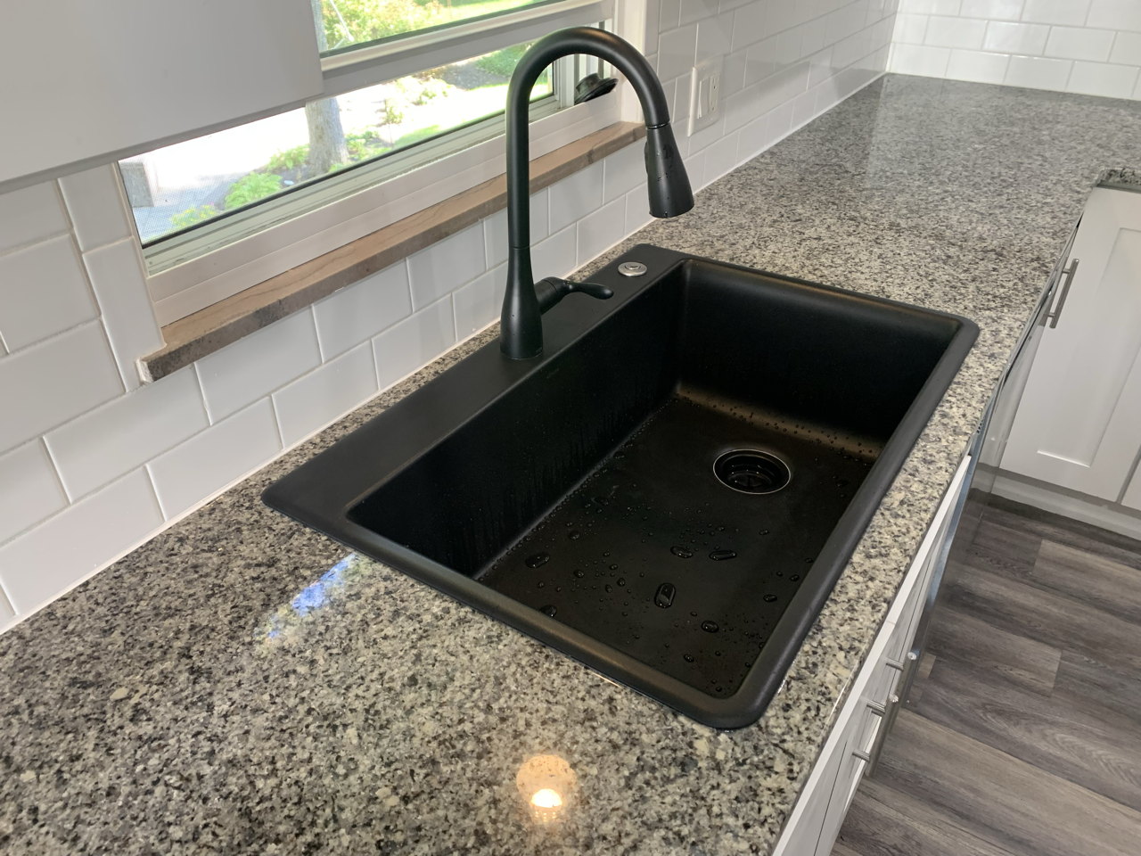 Black kitchen sink installed in a granite countertop with a black faucet, beneath a window with a view of a garden.