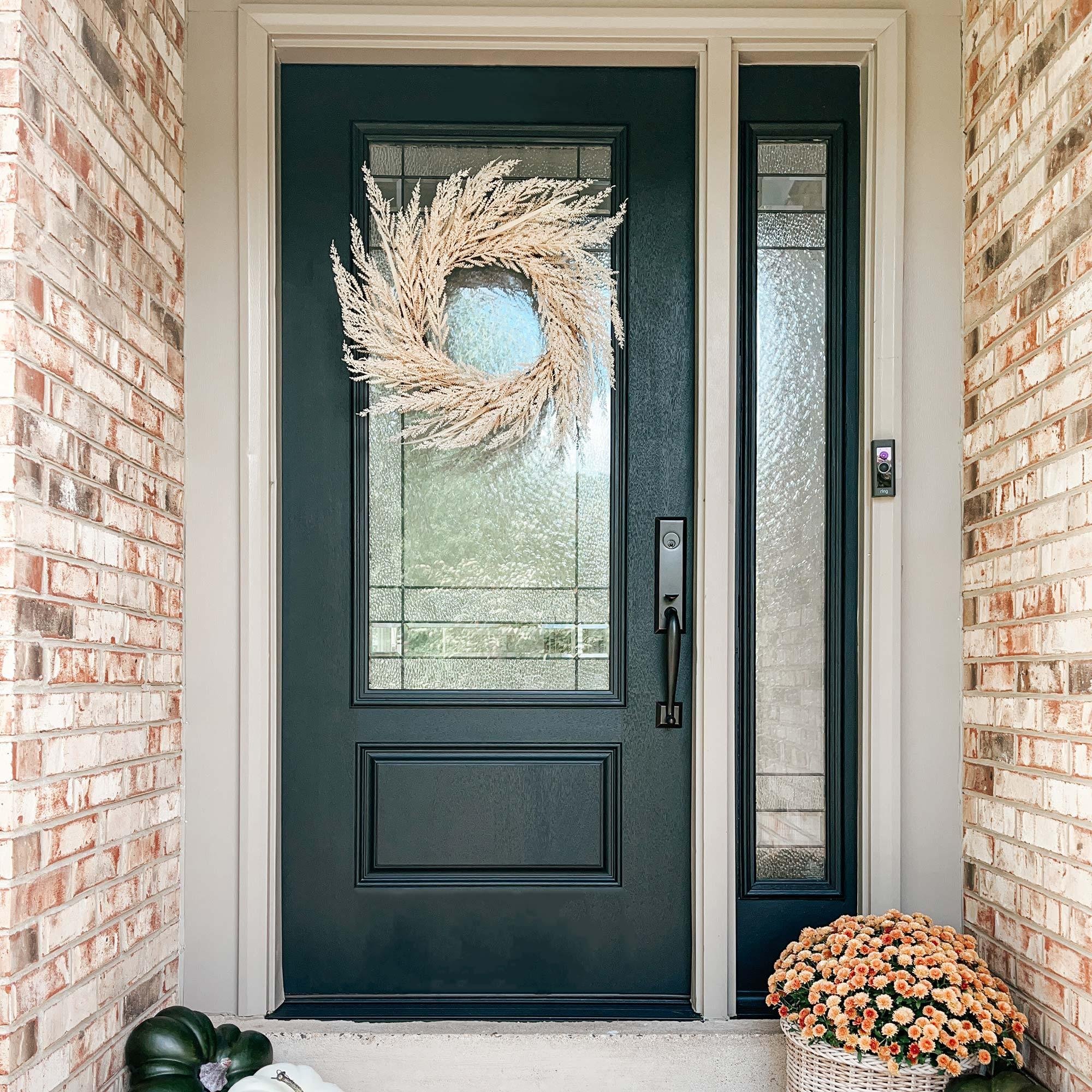 Black front door with glass panels, framed by brick walls on sides, decorated with a white wreath, with potted plants and flowers outside.
