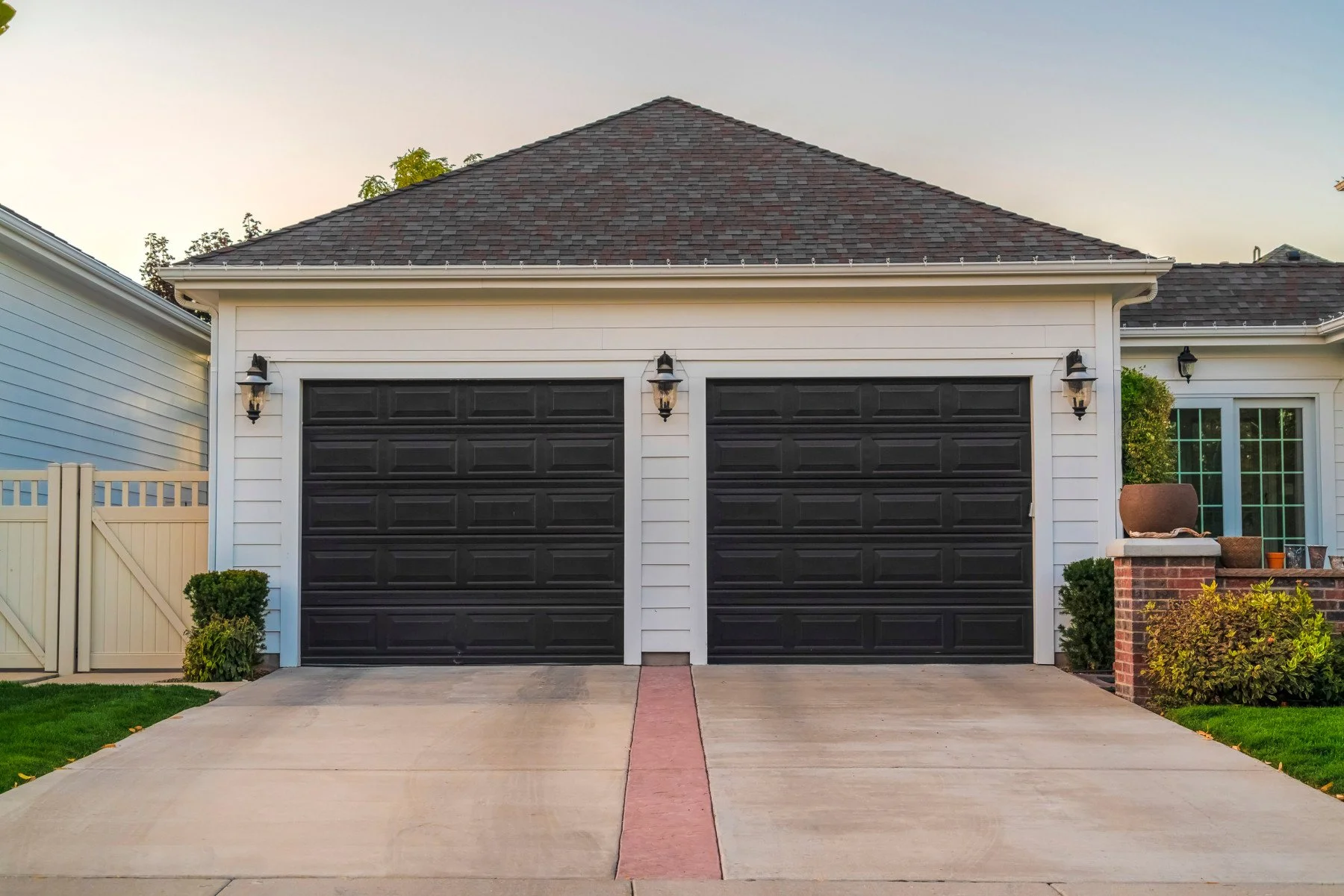 A suburban house with a double garage and black garage doors. The house has white siding, a gray shingled roof, and decorative outdoor lighting. There is a driveway leading to the garage, flanked by green bushes and a brick planter on the right with potted plants. A white fence is on the left side of the driveway.