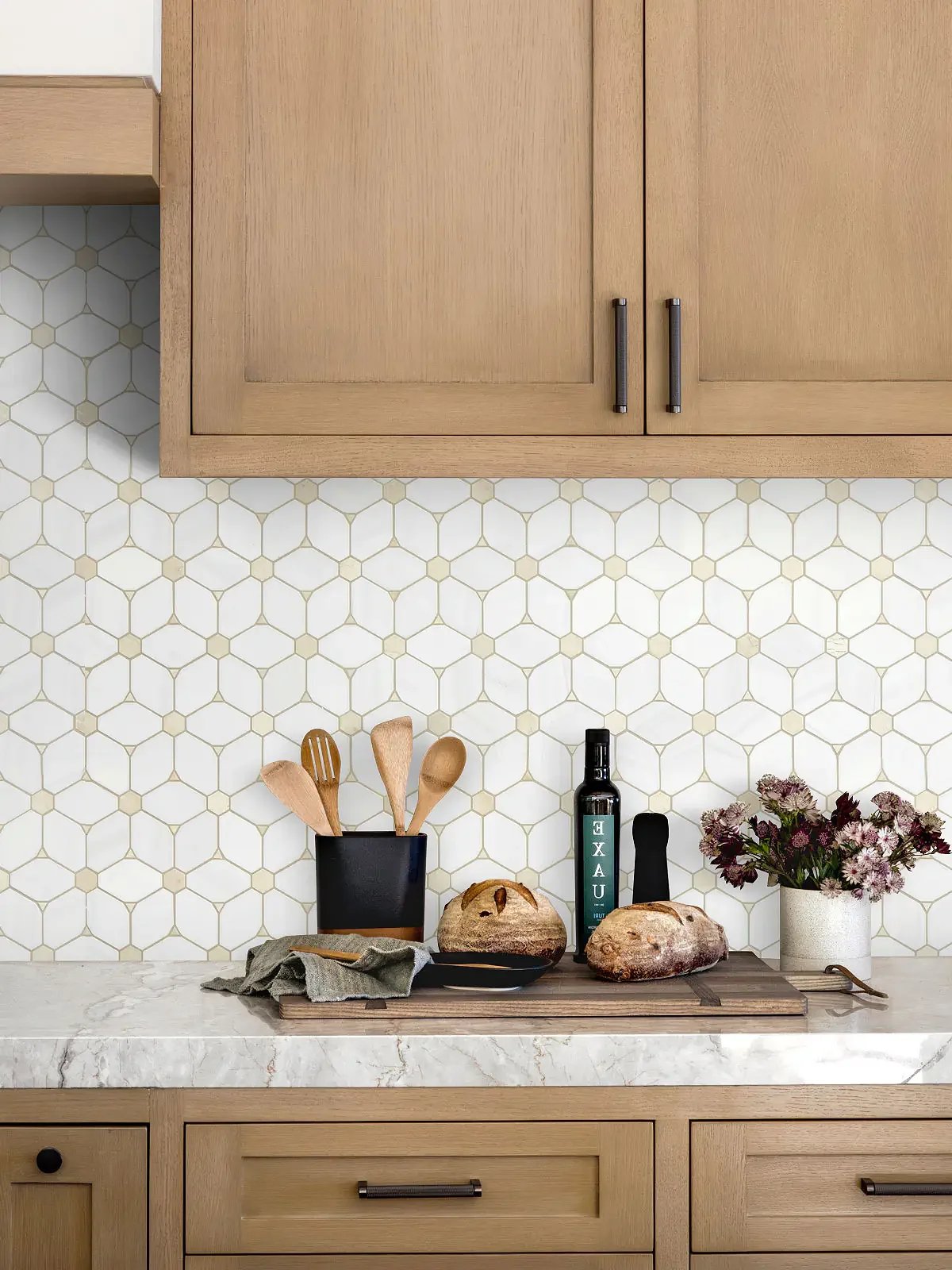 Kitchen countertop with wooden utensils in a black container, two loaves of bread, herbs in a white pot, a bottle of spray, and a cloth, with a honeycomb patterned tile backsplash and wooden cabinets.