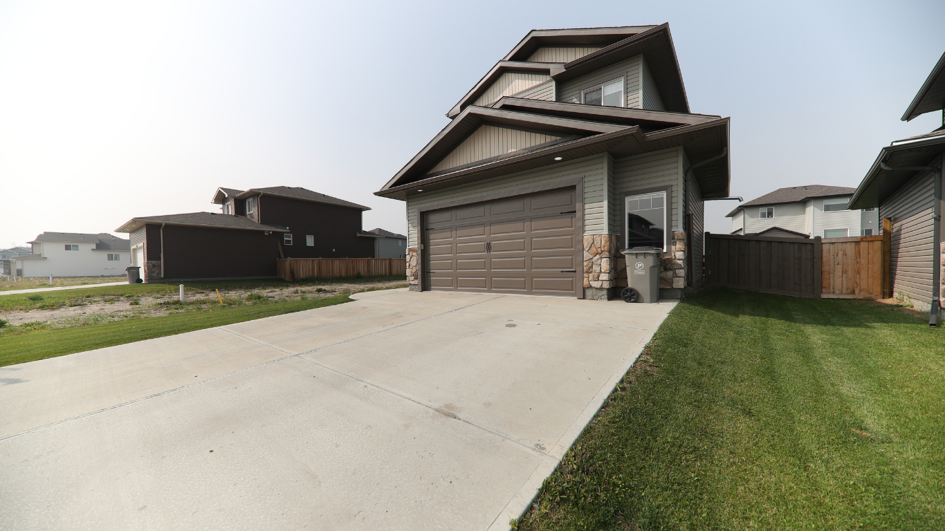 A suburban house with a two-car garage, green siding, stone accents, a driveway, and a small patch of grass beside it. There are neighboring houses visible in the background.