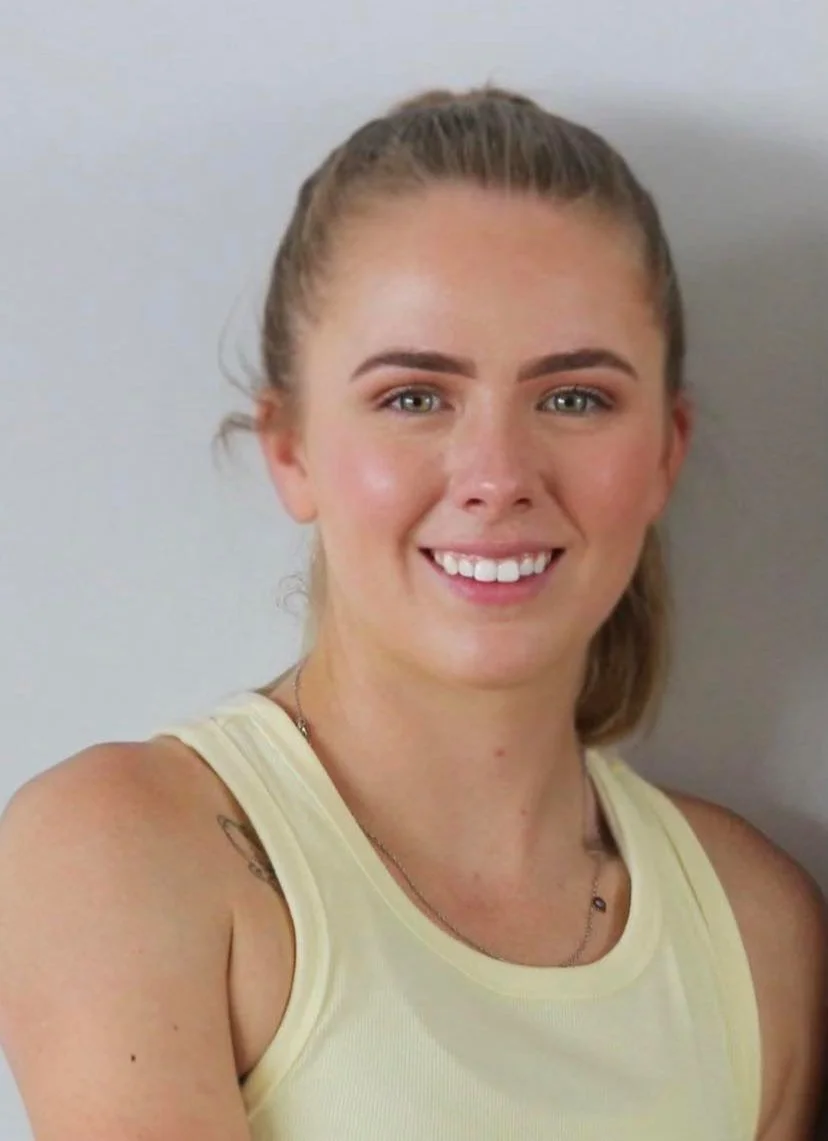 Person smiling at the camera, wearing a yellow tank top, with hair tied back and a tattoo on the shoulder.