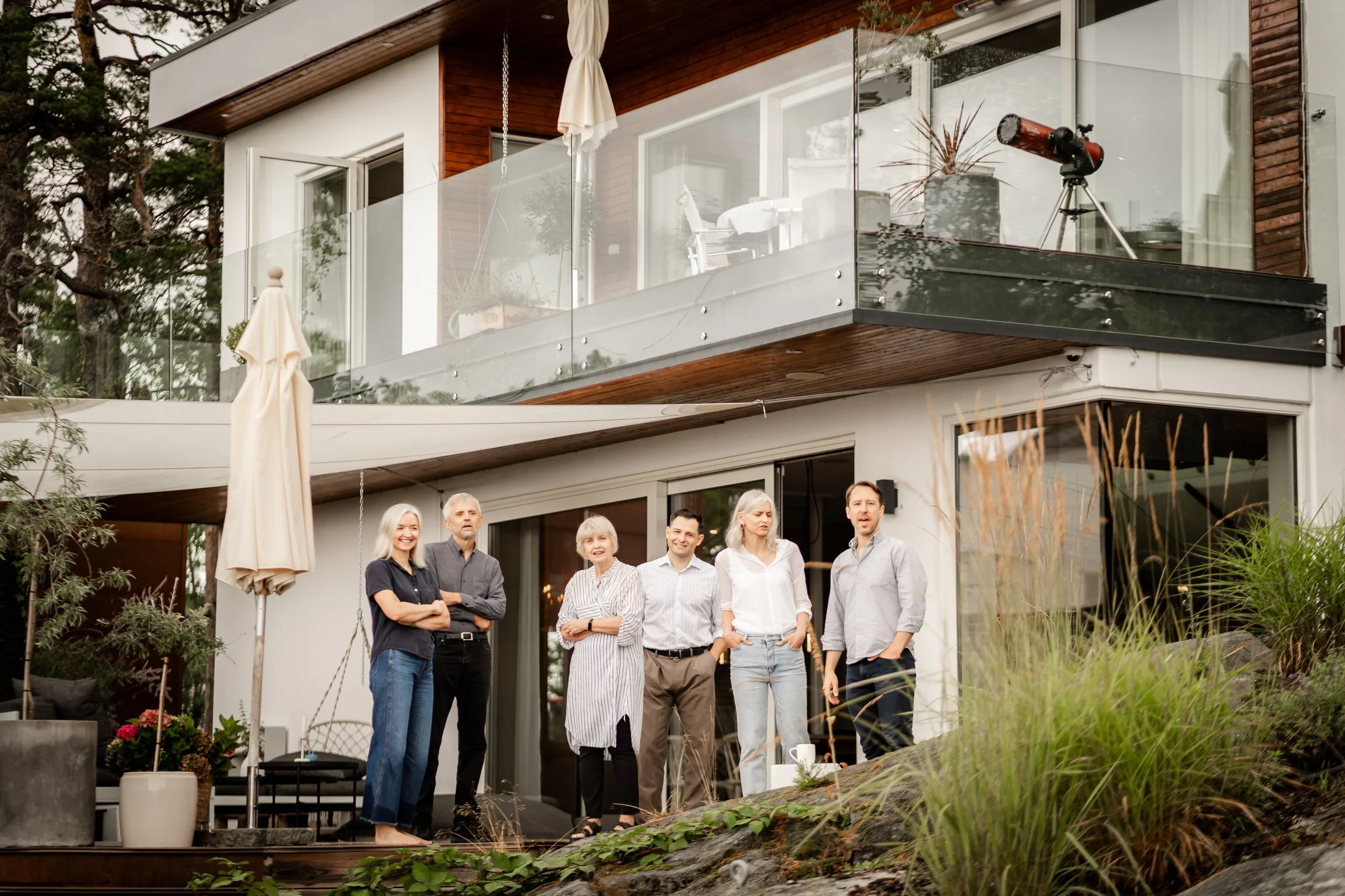 Group of six people standing outside a modern multi-story house with large glass sliding doors and balcony, surrounded by greenery.