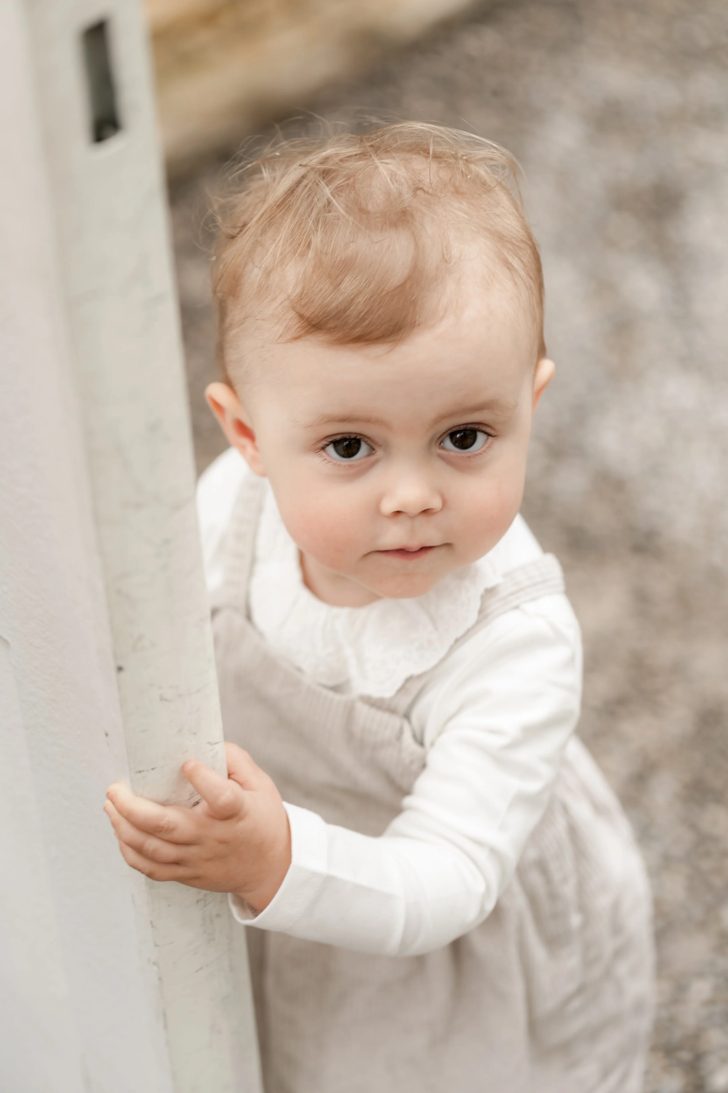 Young child with light hair and brown eyes peeking around a white wall, wearing a cream-colored dress with a white long-sleeve shirt underneath.