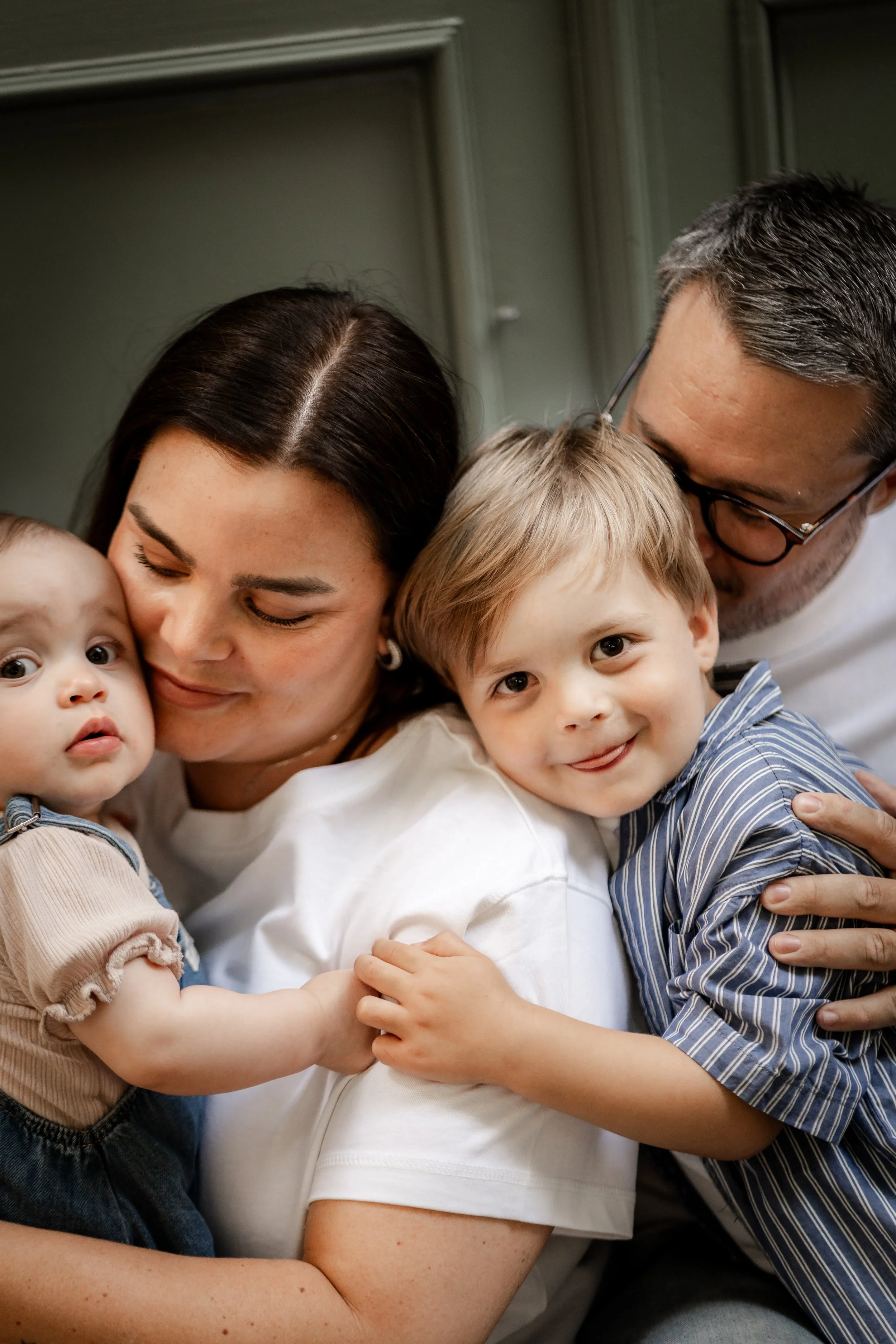 A family of four hugging inside a house, including a woman, a man, a young boy, and a baby.