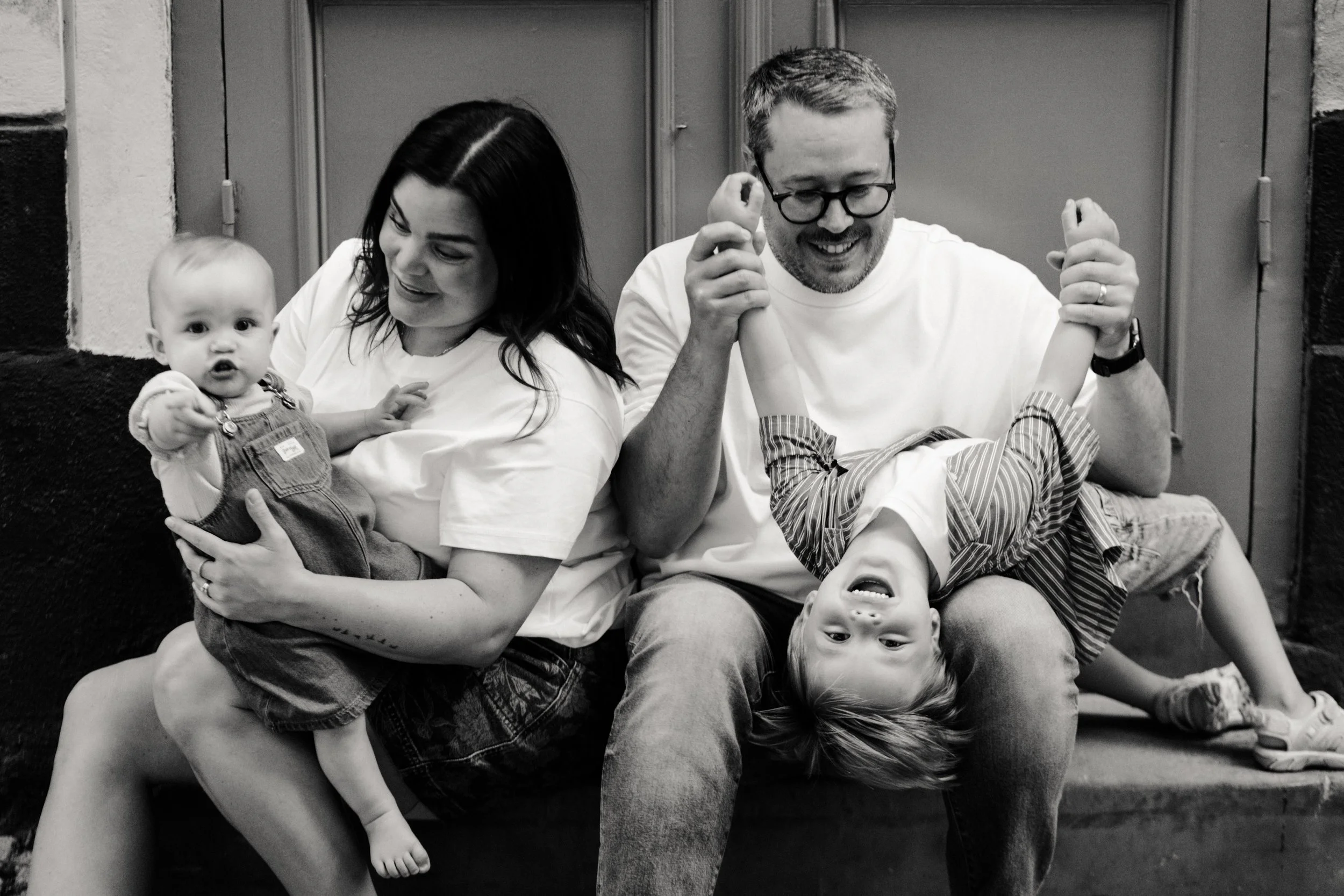 A black-and-white photo of a family sitting on a bench. The woman is holding a baby girl, and a man is playing with a little boy upside down on his lap. All are smiling and appear to be having fun.
