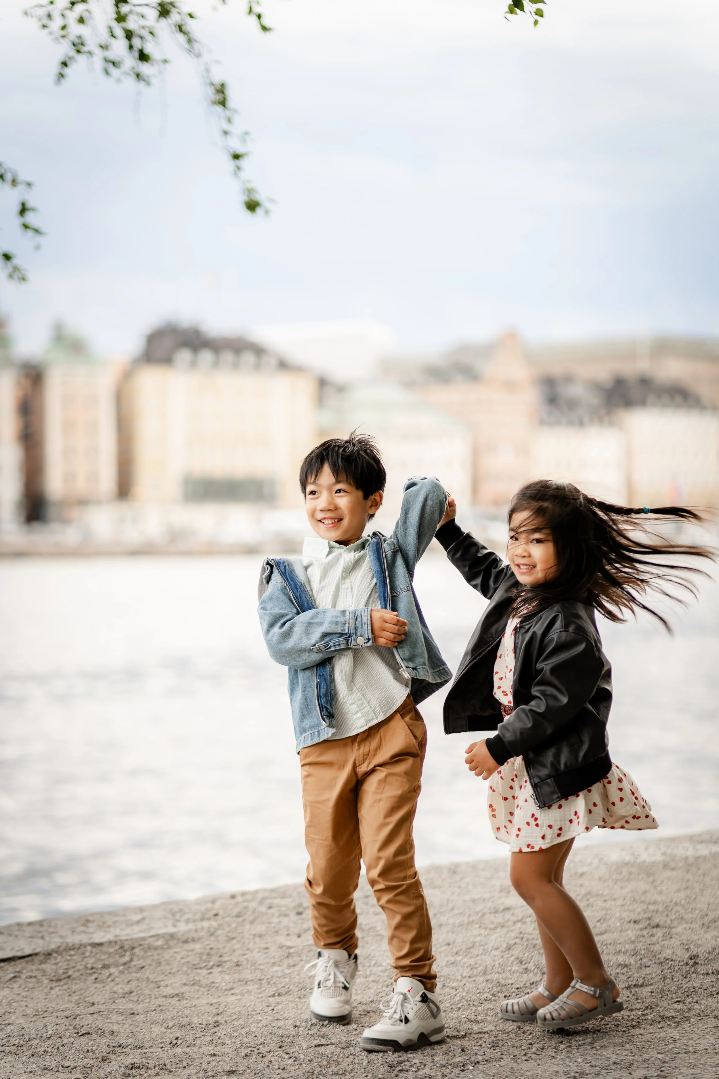 Two children, a boy and a girl, are playing by the water on a cloudy day, smiling and holding hands.