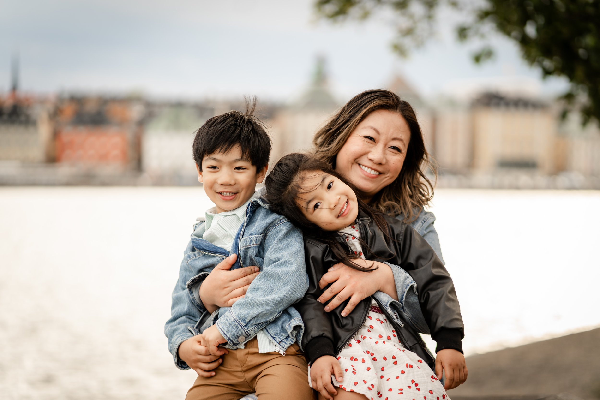A woman hugging two children outdoors near a body of water with city buildings and trees in the background.