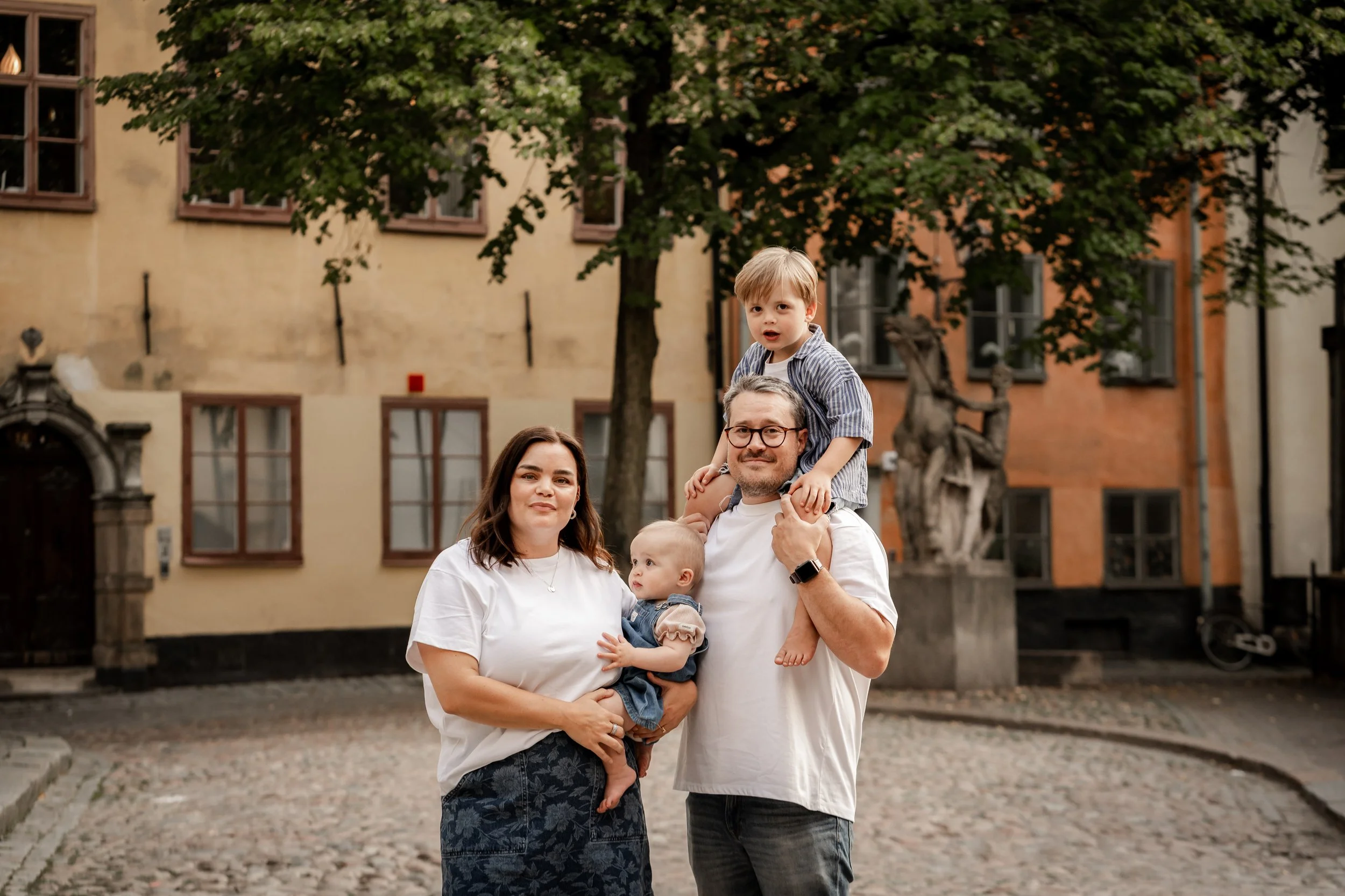 A family of four standing outdoors in front of a yellow building, with a large tree behind them. The father is holding a young toddler on his shoulders, and the mother is holding a baby girl. All are smiling and looking at the camera.