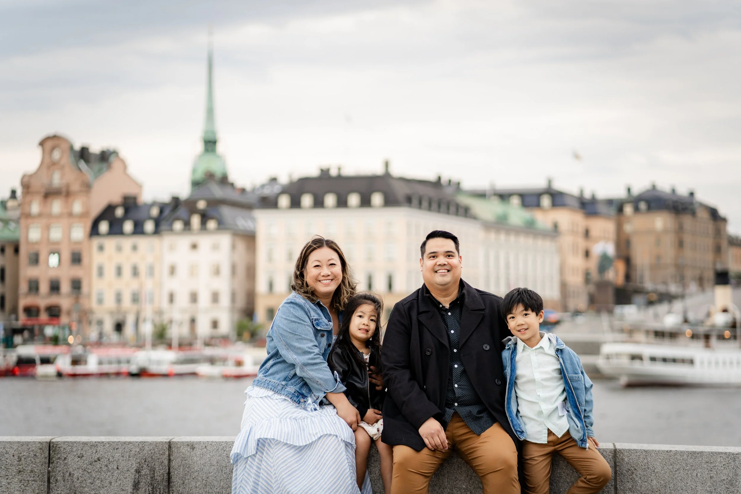 A family of four sitting on a stone wall in front of a cityscape with boats on water and historic buildings, likely in Europe. The family includes a woman, a man, a young girl, and a young boy, all smiling.