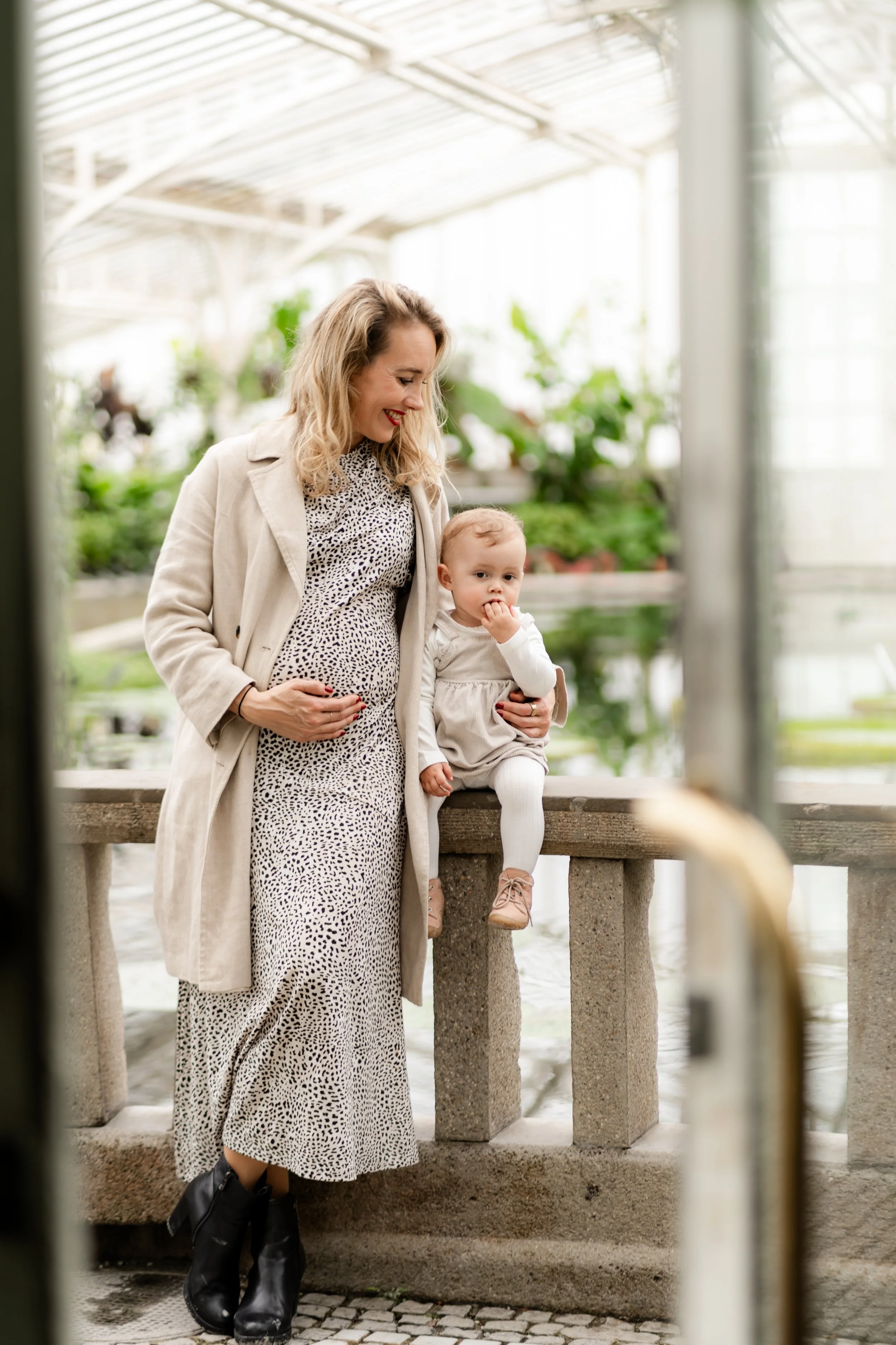 A woman with blonde curly hair in a leopard print dress and beige coat holds a young girl in a white dress and tights by a stone railing in a greenhouse or botanical garden.