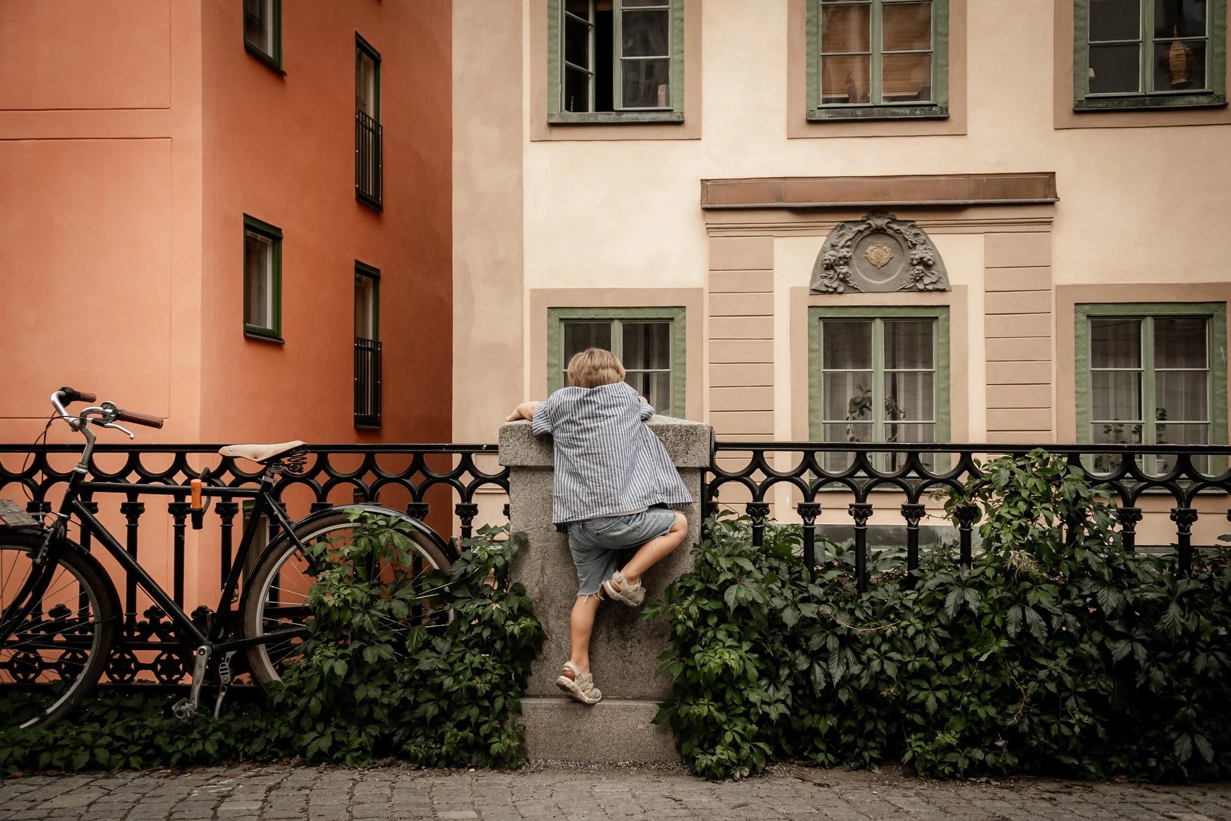 A young boy in a striped shirt and denim shorts climbing over a low stone wall, leaning on a marble post, with a bicycle parked nearby and greenery in the foreground. In the background are multi-story buildings with European-style architecture and pa