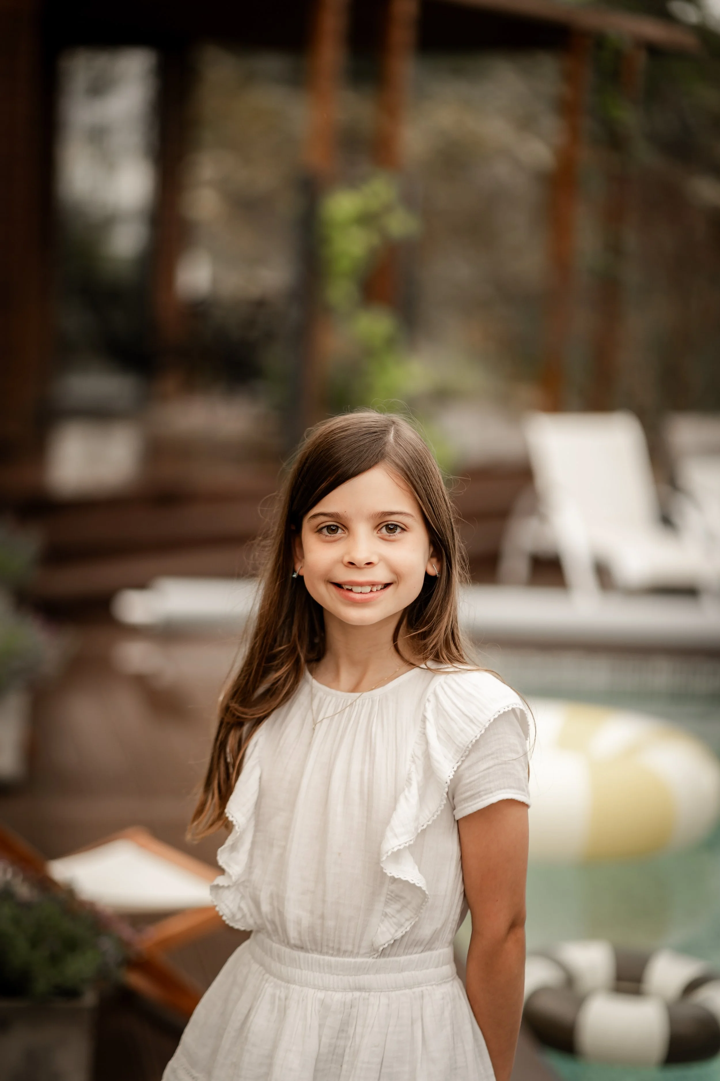 A smiling young girl with long brown hair standing outdoors near a wooden deck and pool, with lounge chairs and a water inflatable in the background.