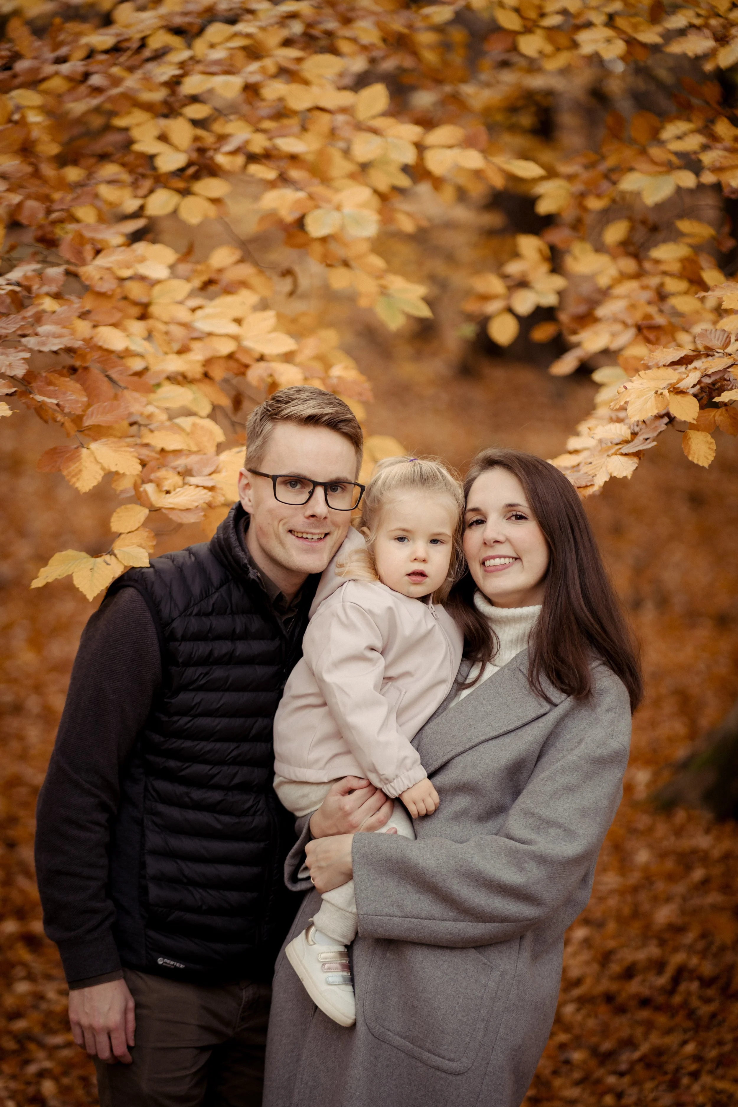 A family of three stands together in a park surrounded by autumn leaves. A man with glasses, a woman with long dark hair, and a young girl with blonde hair pose for a photo.