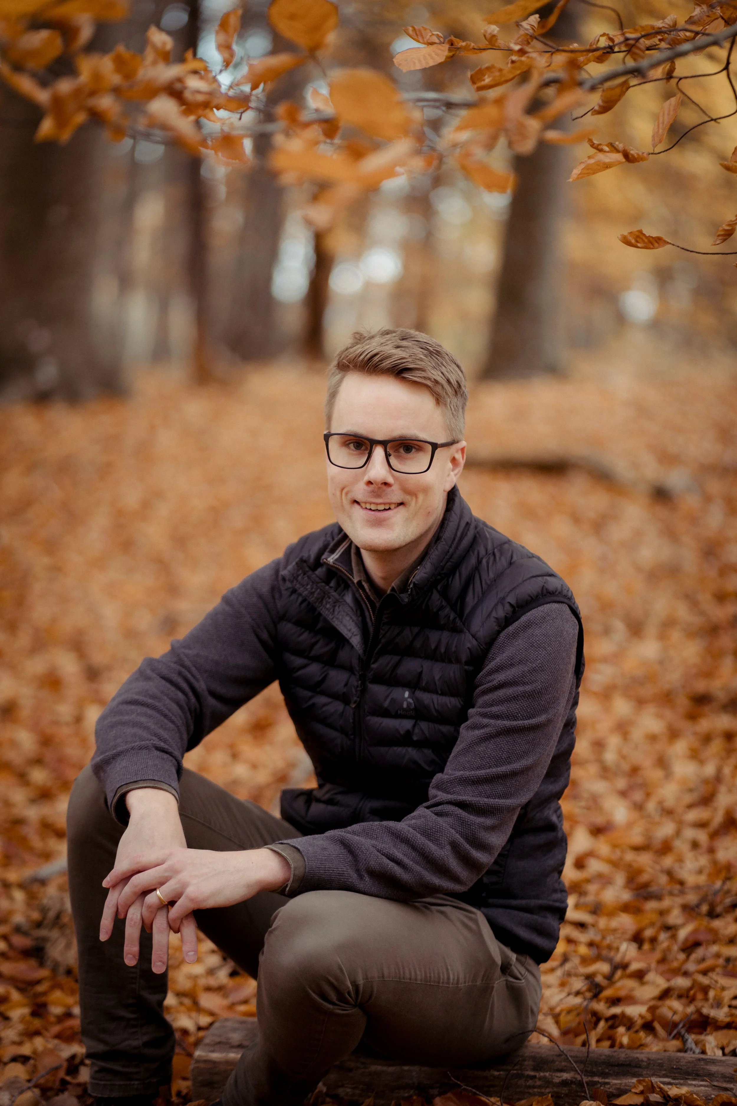 A young man with glasses, wearing a black vest and gray sweater, sitting on a log in a forest during autumn with fallen leaves on the ground.
