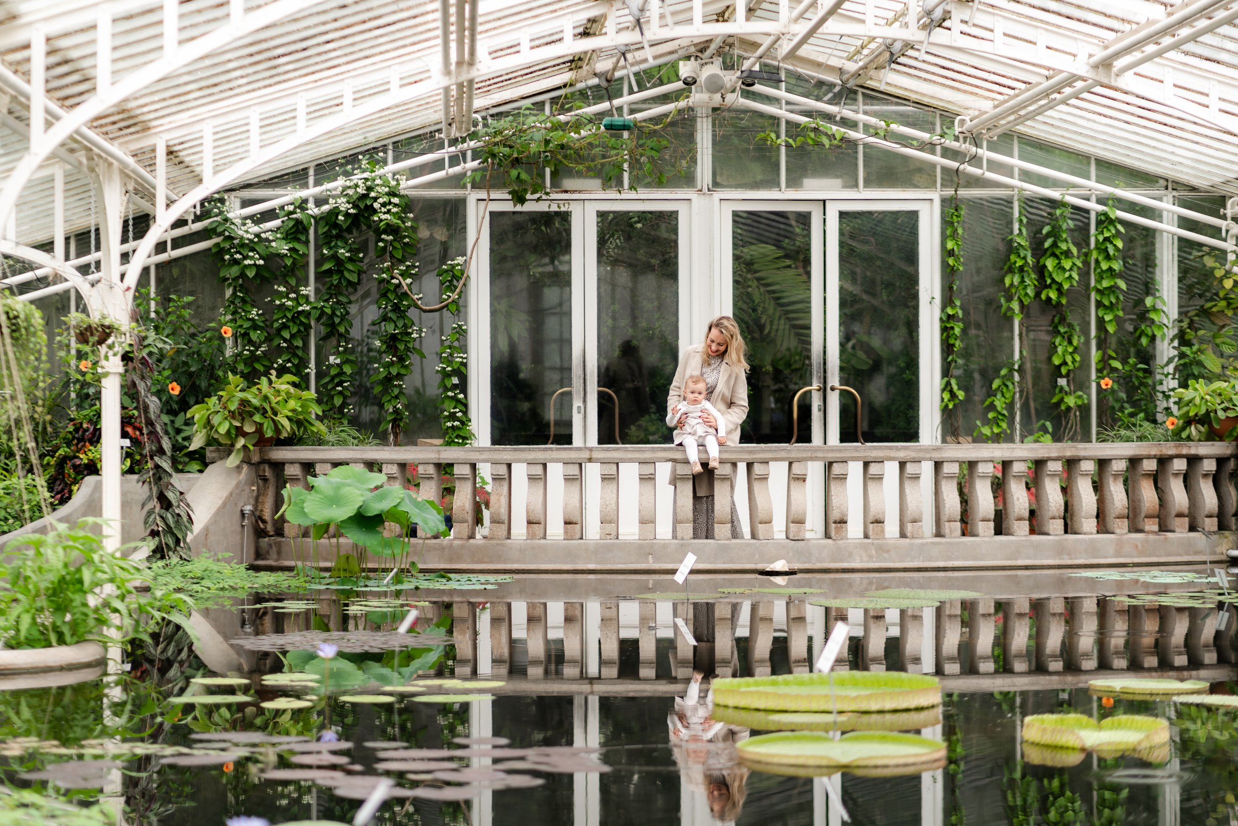 A woman and a baby sitting on a railing in a greenhouse with lush green plants and a pond in front.