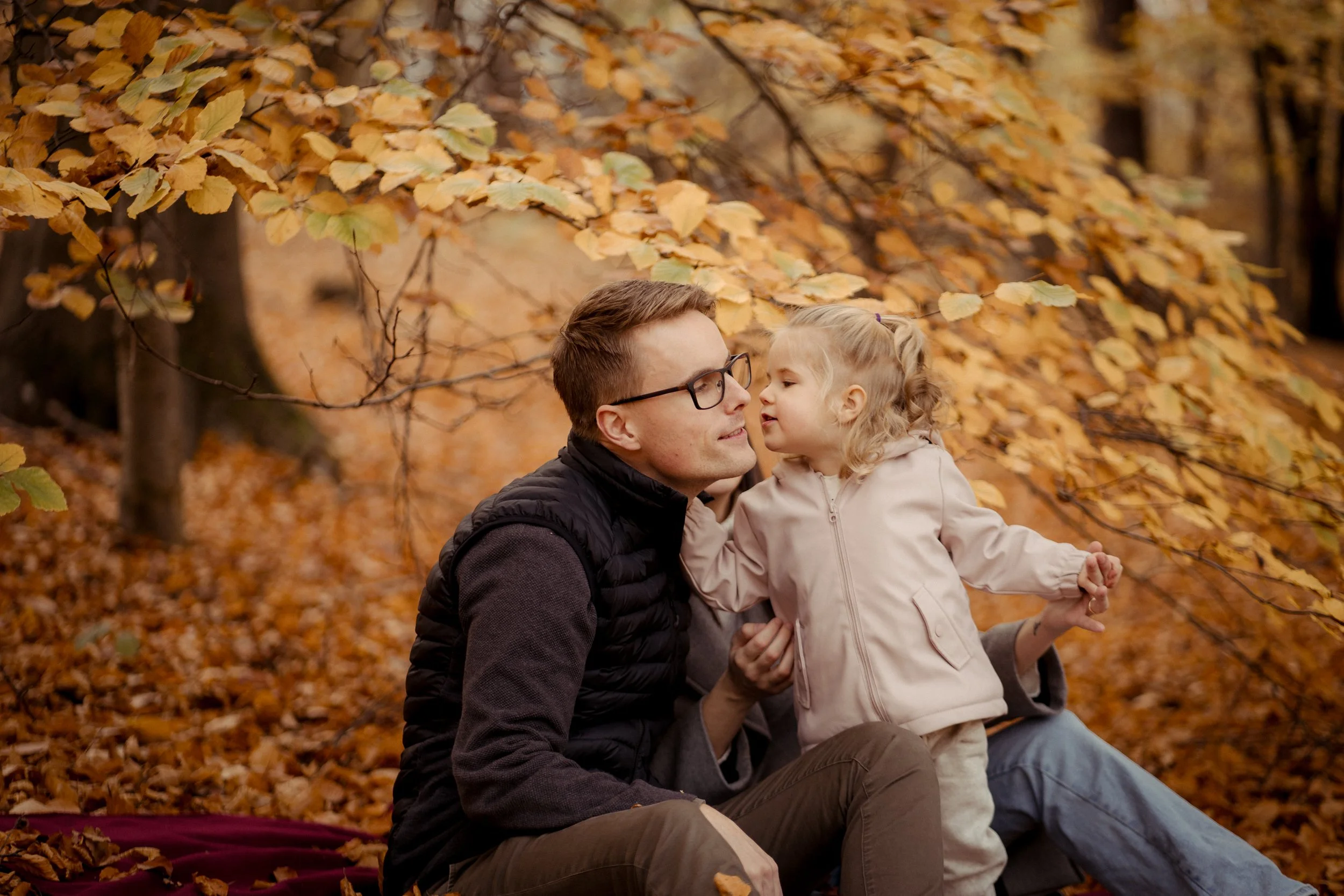 A father and daughter sit on a blanket in a fall forest, with leaves in shades of yellow and orange surrounding them. The daughter leans toward her father, whispering or speaking closely.