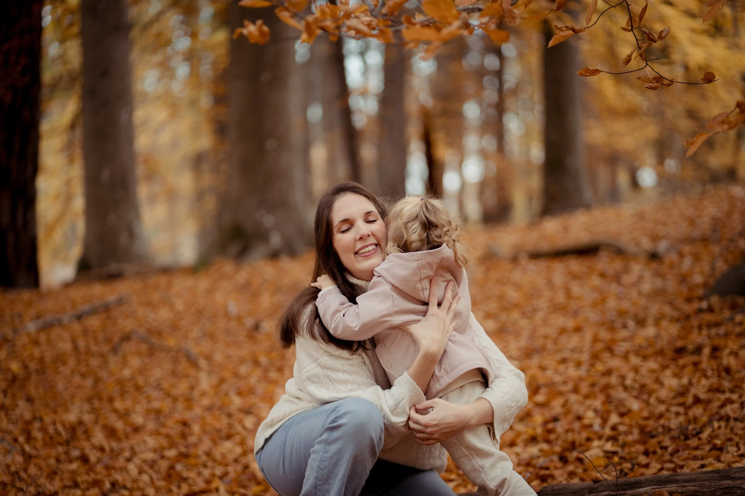 A woman and a young girl, possibly mother and daughter, hugging and smiling in an autumn forest with fallen leaves and tall trees.