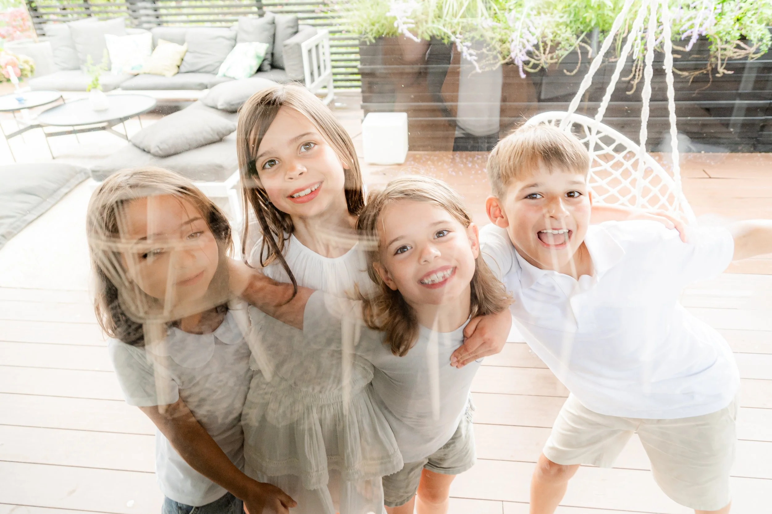 Four children peering through glass door, smiling and posing with their arms around each other indoors with a living room in the background.