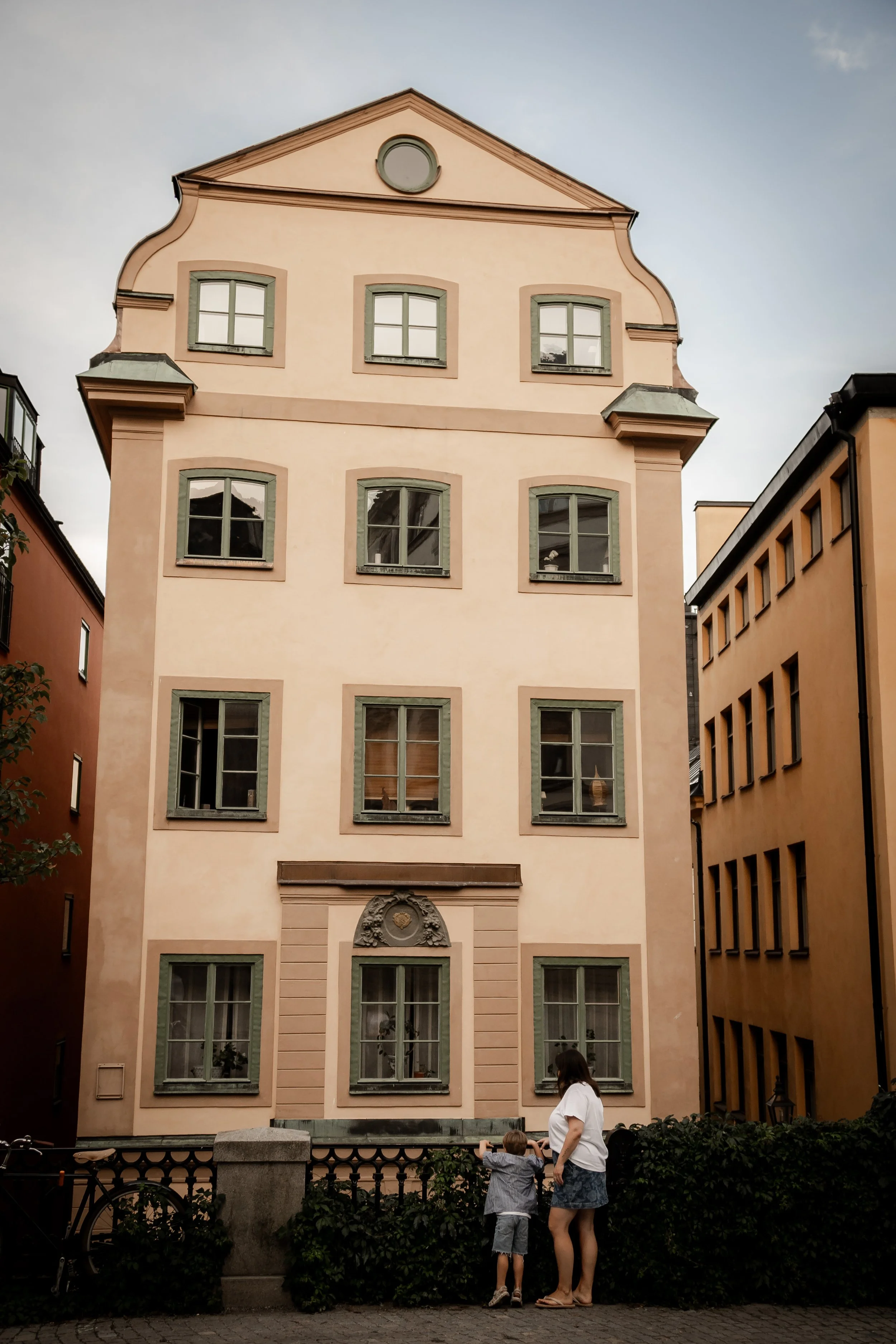 A tall, pale pink, multi-story building with green window frames, decorative architectural details, and a clock circle near the top. In front, a woman and two children stand by a black fence and look at the building.
