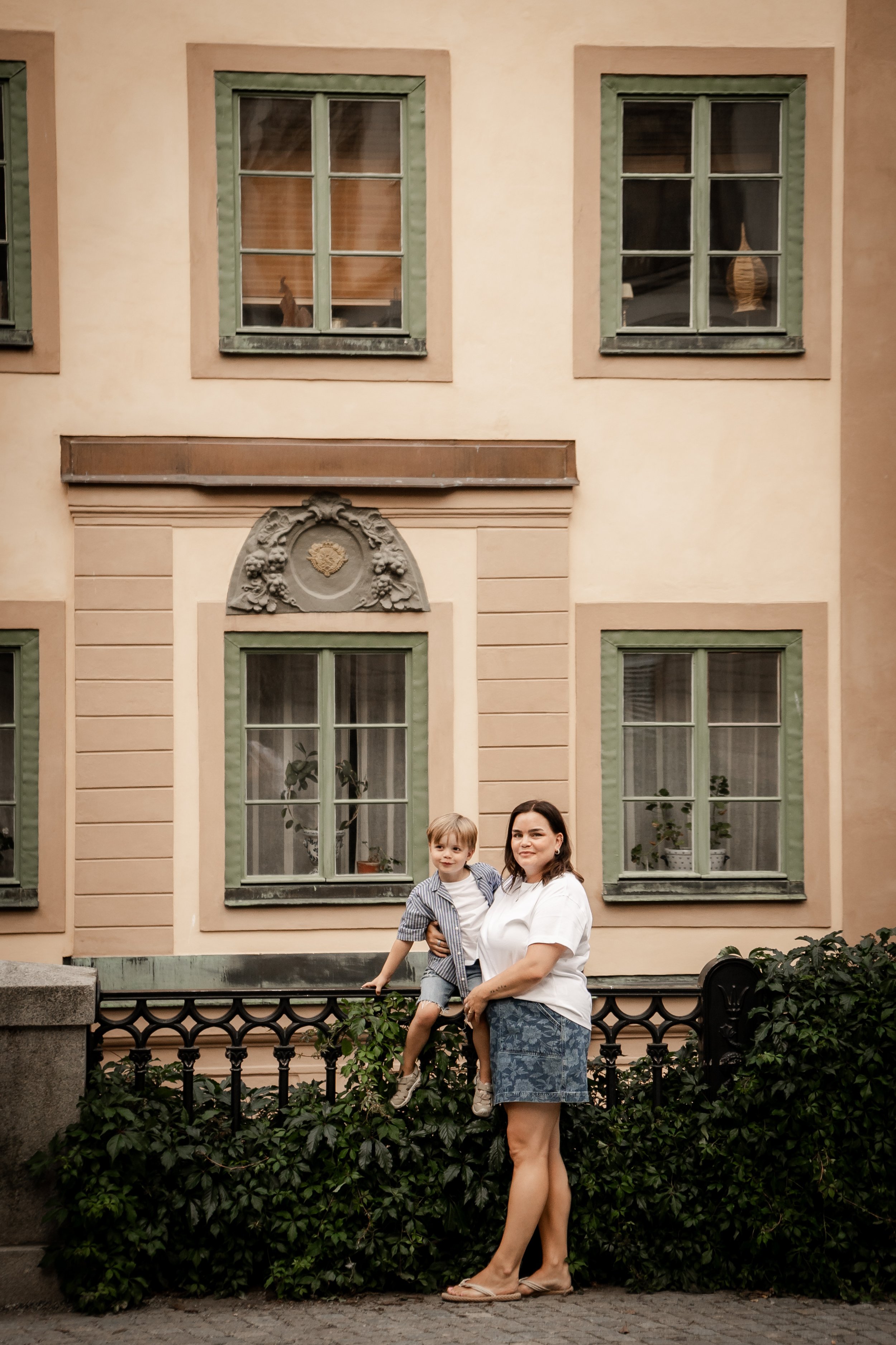 A woman and a young boy standing outdoors in front of a house with cream-colored walls, green window frames, and decorative details.