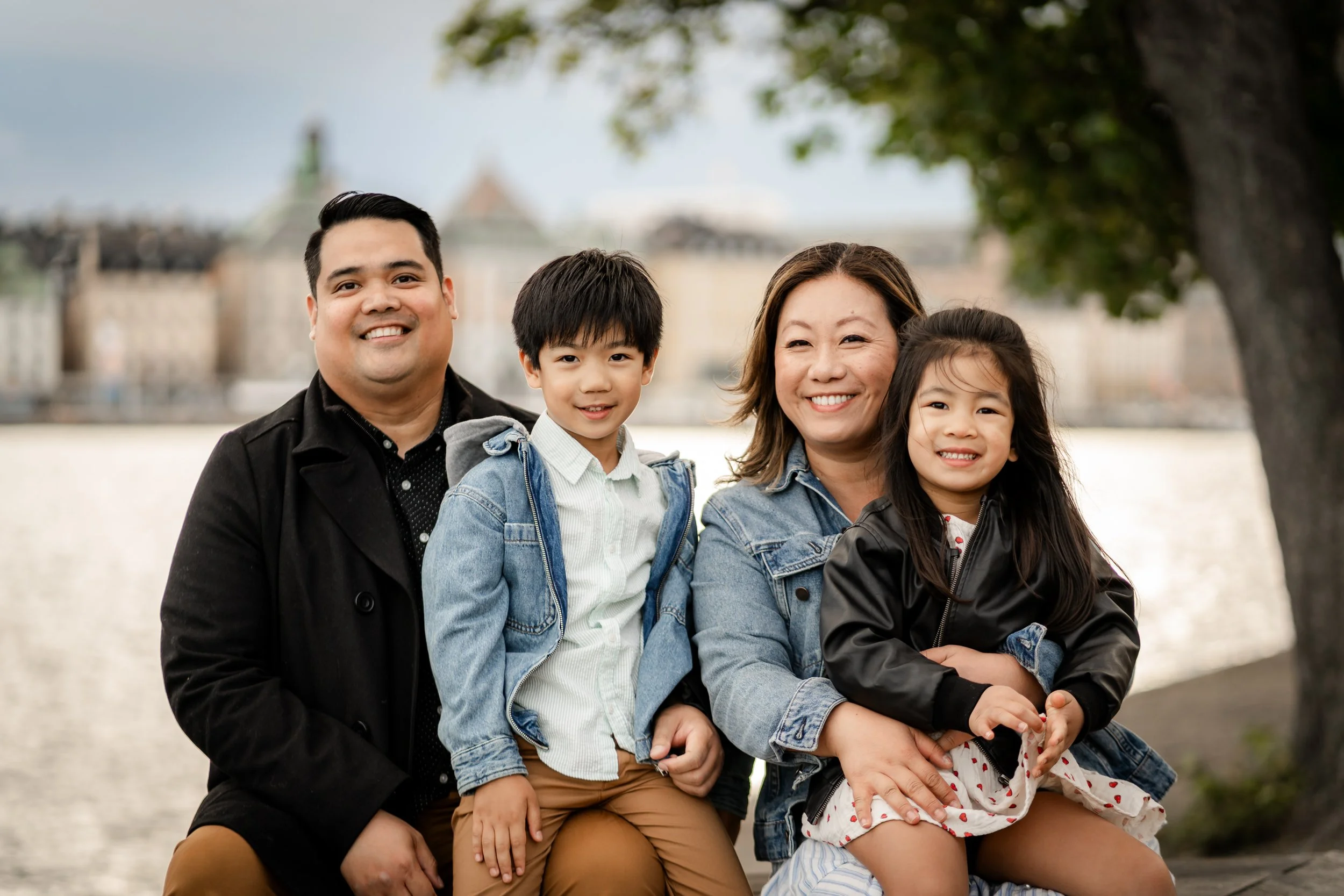 Family of four outdoors near water with cityscape in background, smiling.