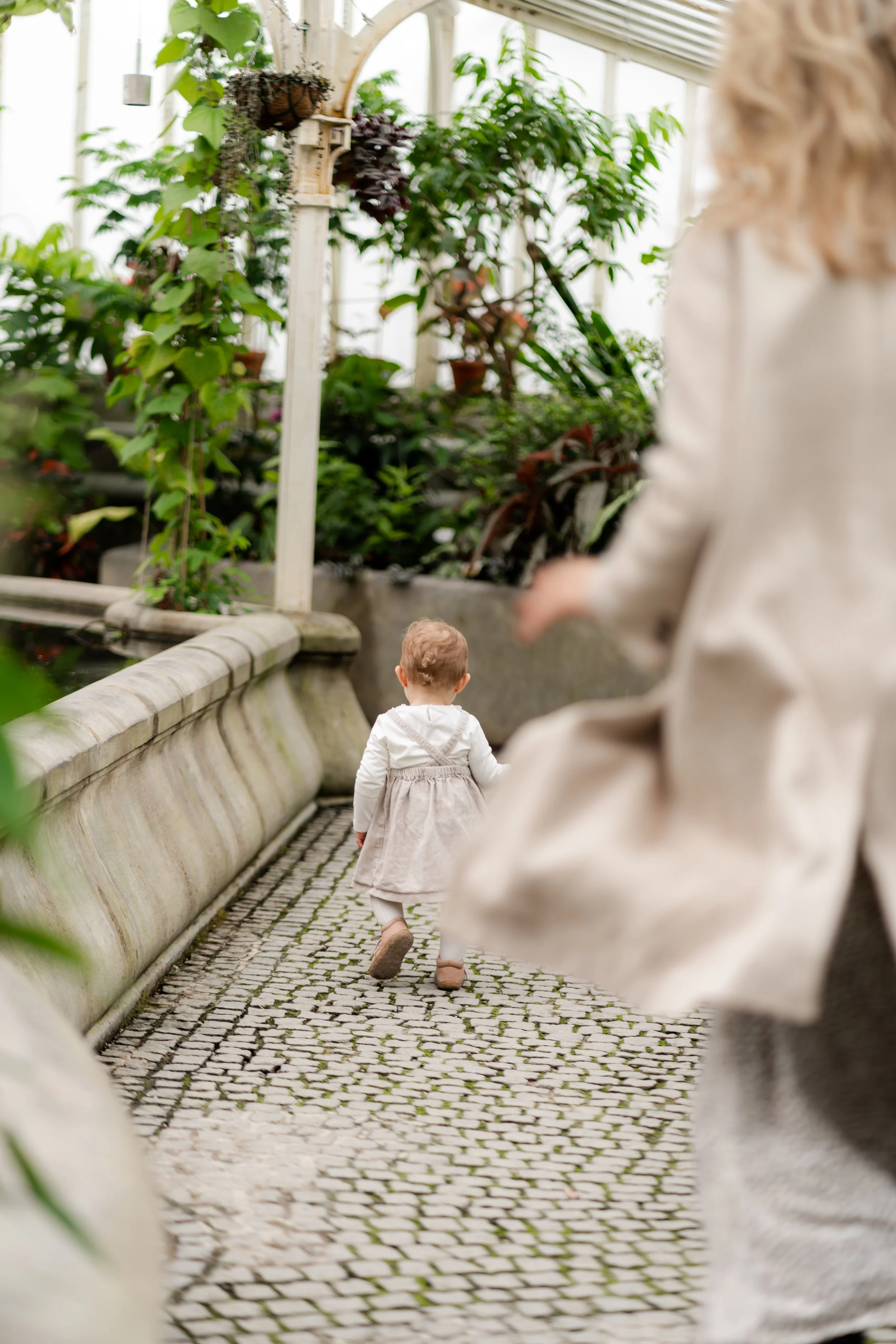 A young child walking down a stone pathway in a greenhouse, with an adult nearby