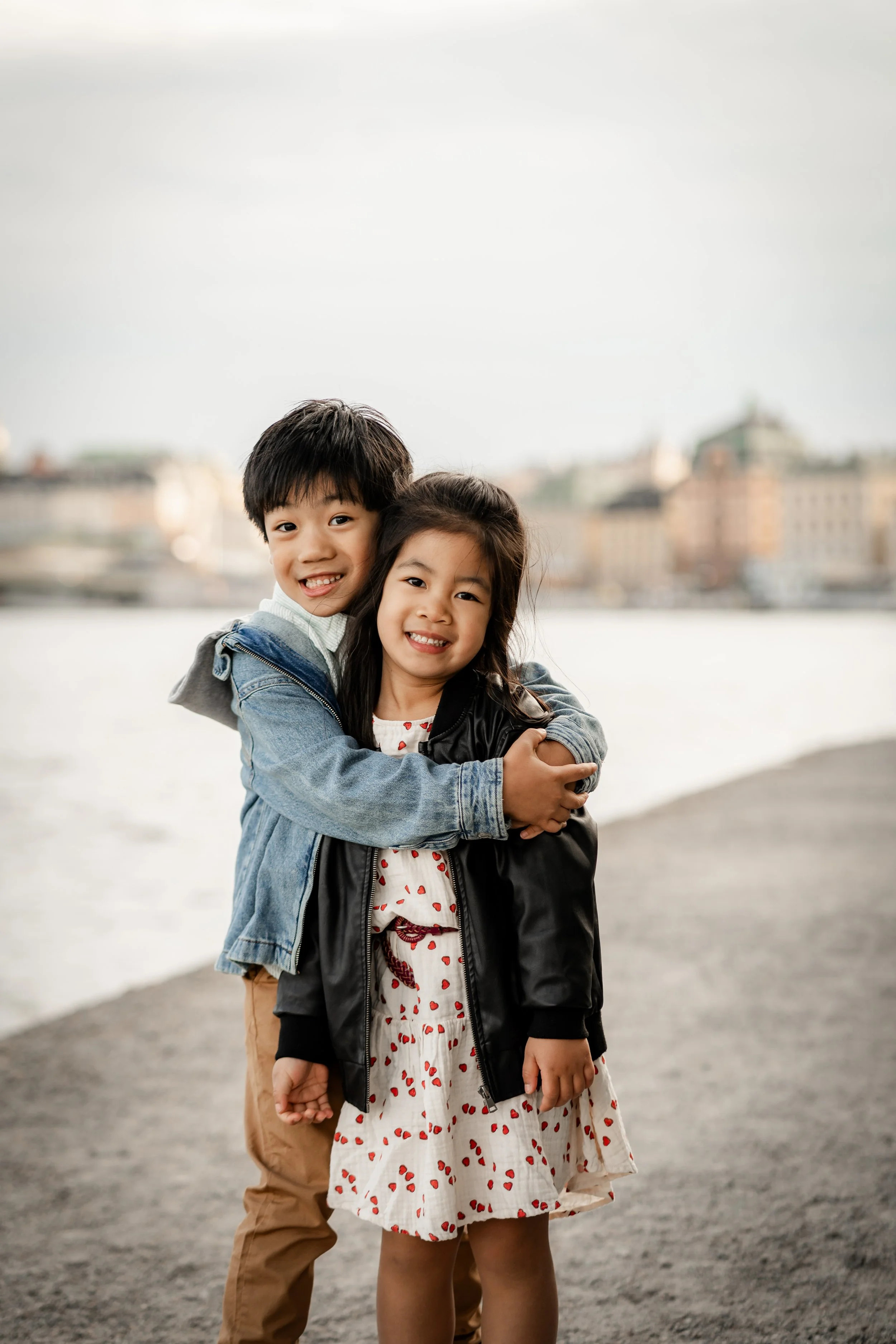 Two children hugging by a river with city buildings in the background.