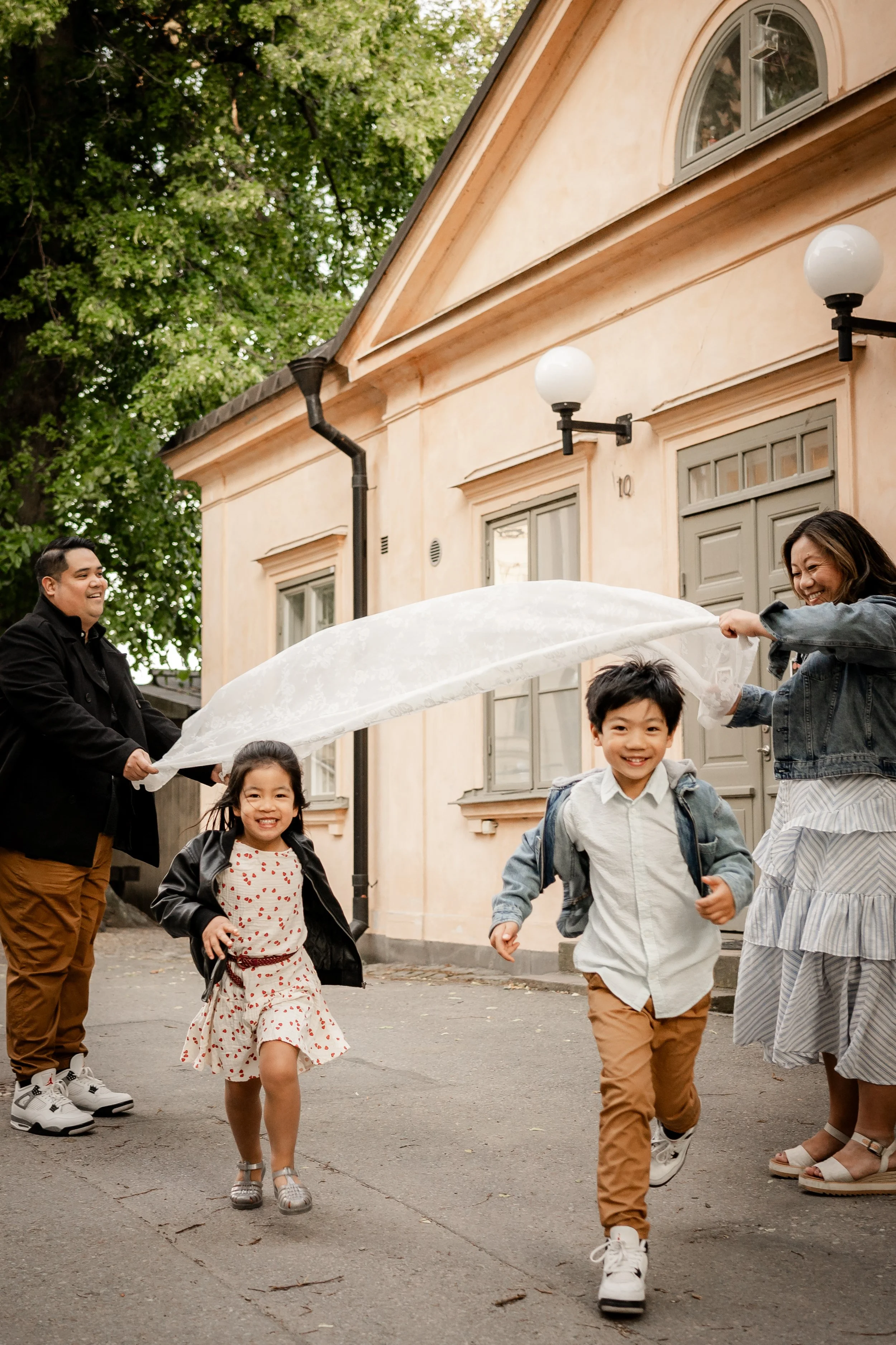 A family of four, including two children and two adults, playfully running outside a house with light-colored walls and large windows. The children are smiling and running toward the camera, while the adults are waving a white cloth or sheet over the