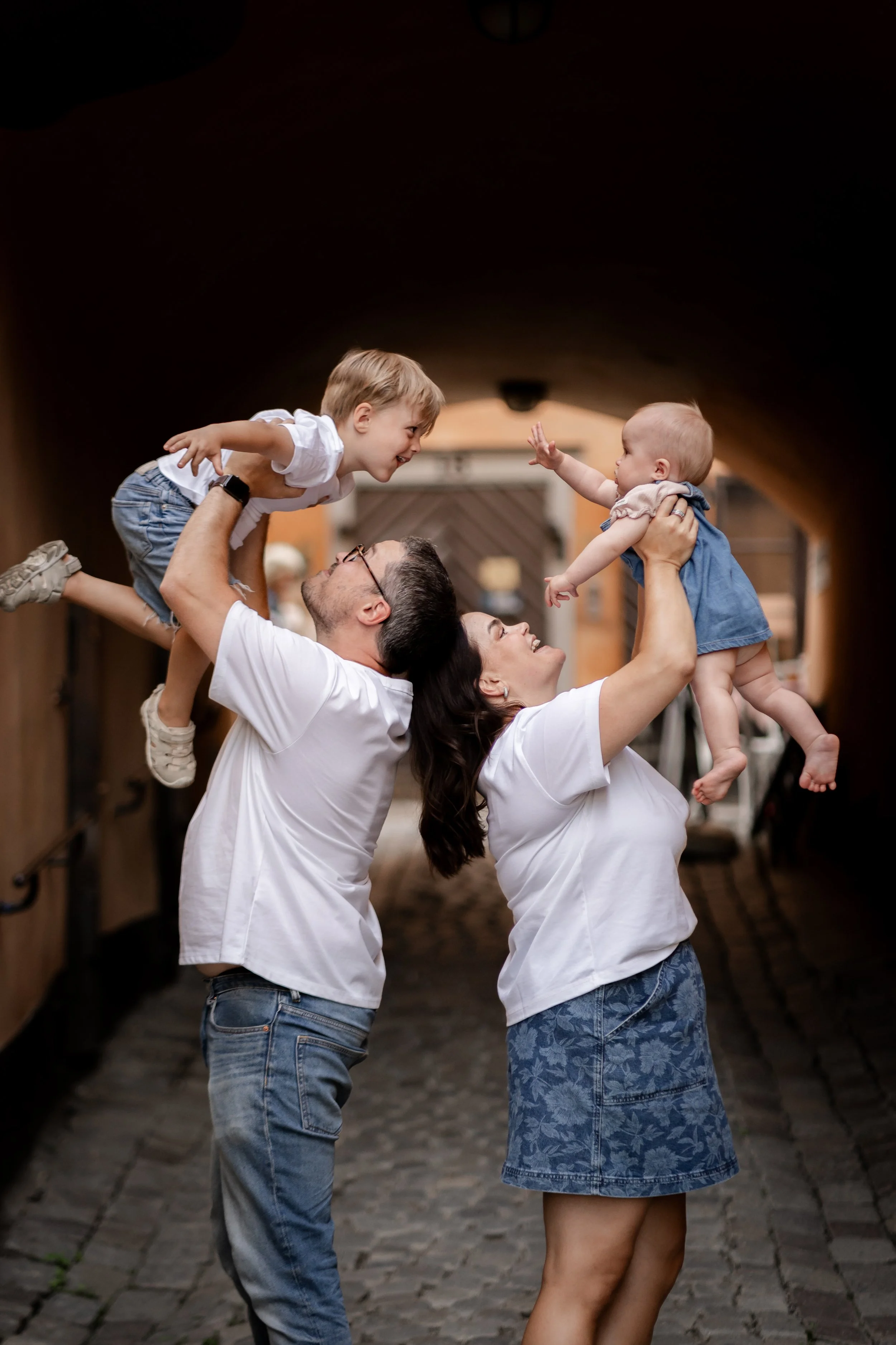Family of four, two parents and two young children, playfully lifting children in an alleyway.