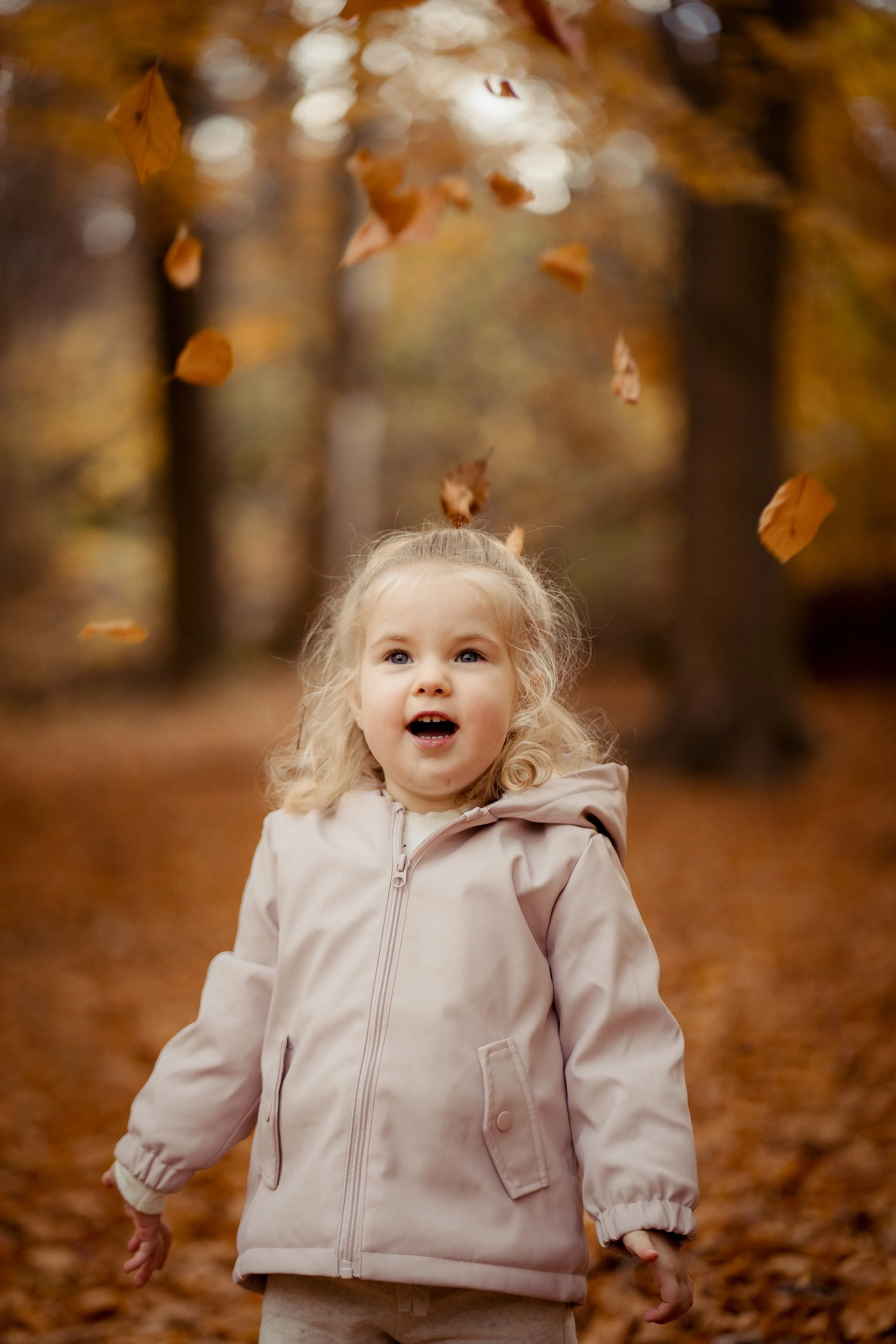 A young girl with blonde, curly hair in a beige jacket is playing in a forest during autumn, with leaves falling around her.