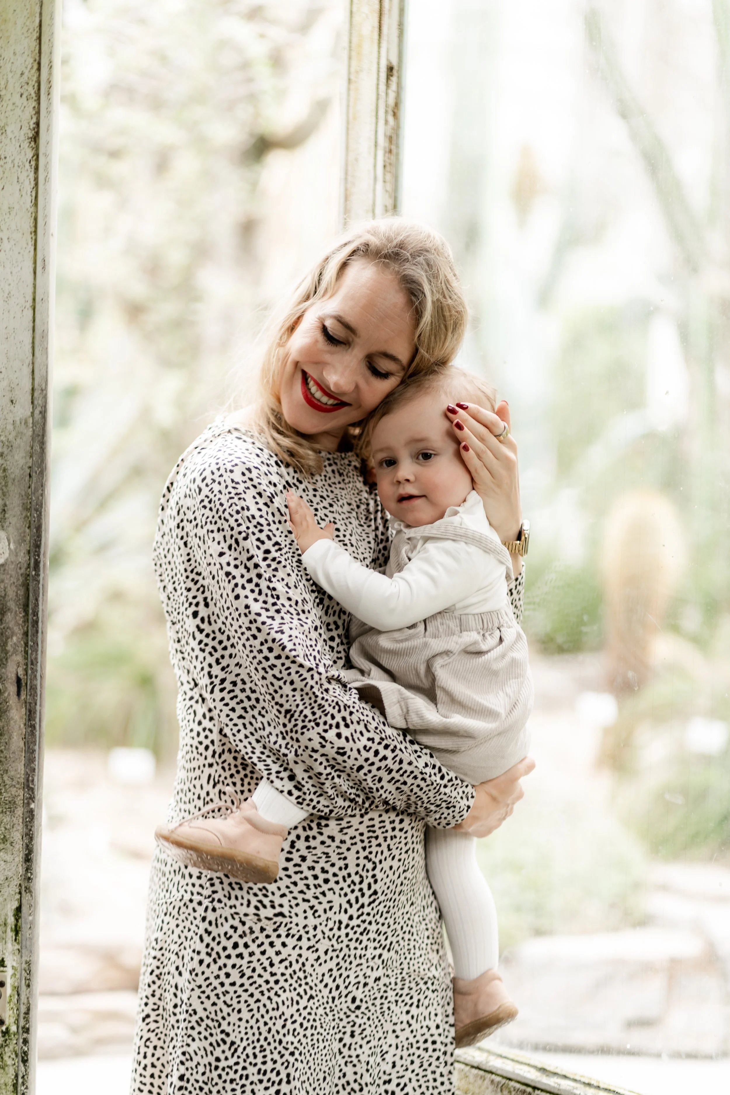A woman with blonde, curly hair and red lipstick holding a young child, both smiling, near a window with a blurred outdoor background.
