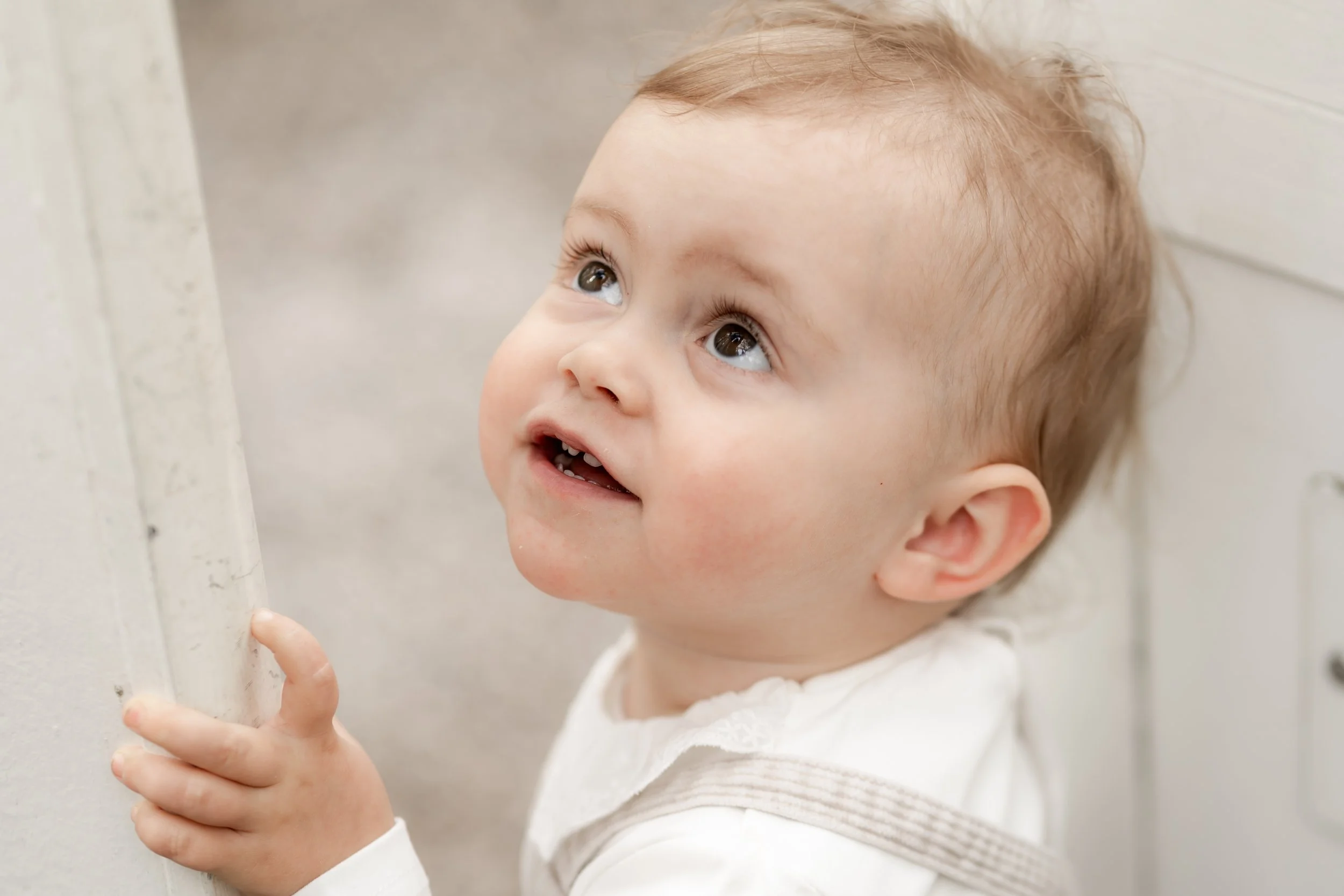 A young child with light brown hair and big eyes looking upward, holding onto a white wall with their right hand.