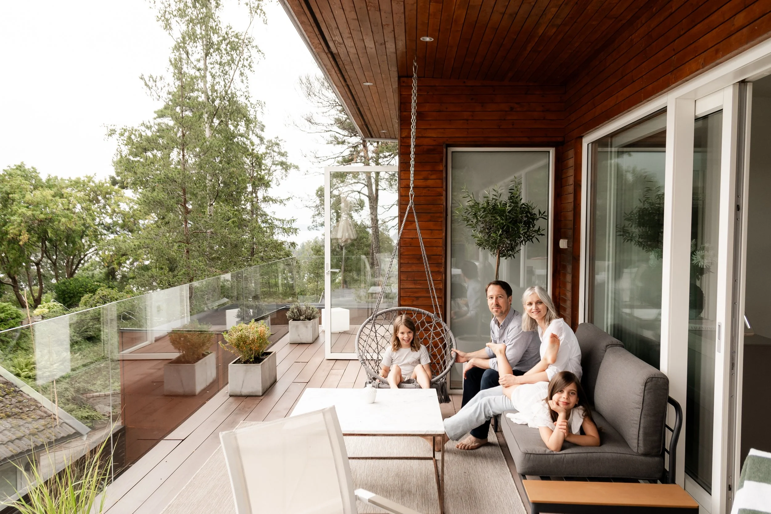 Family of four relaxing on a balcony with glass railing, outdoor plants, and trees in the background.