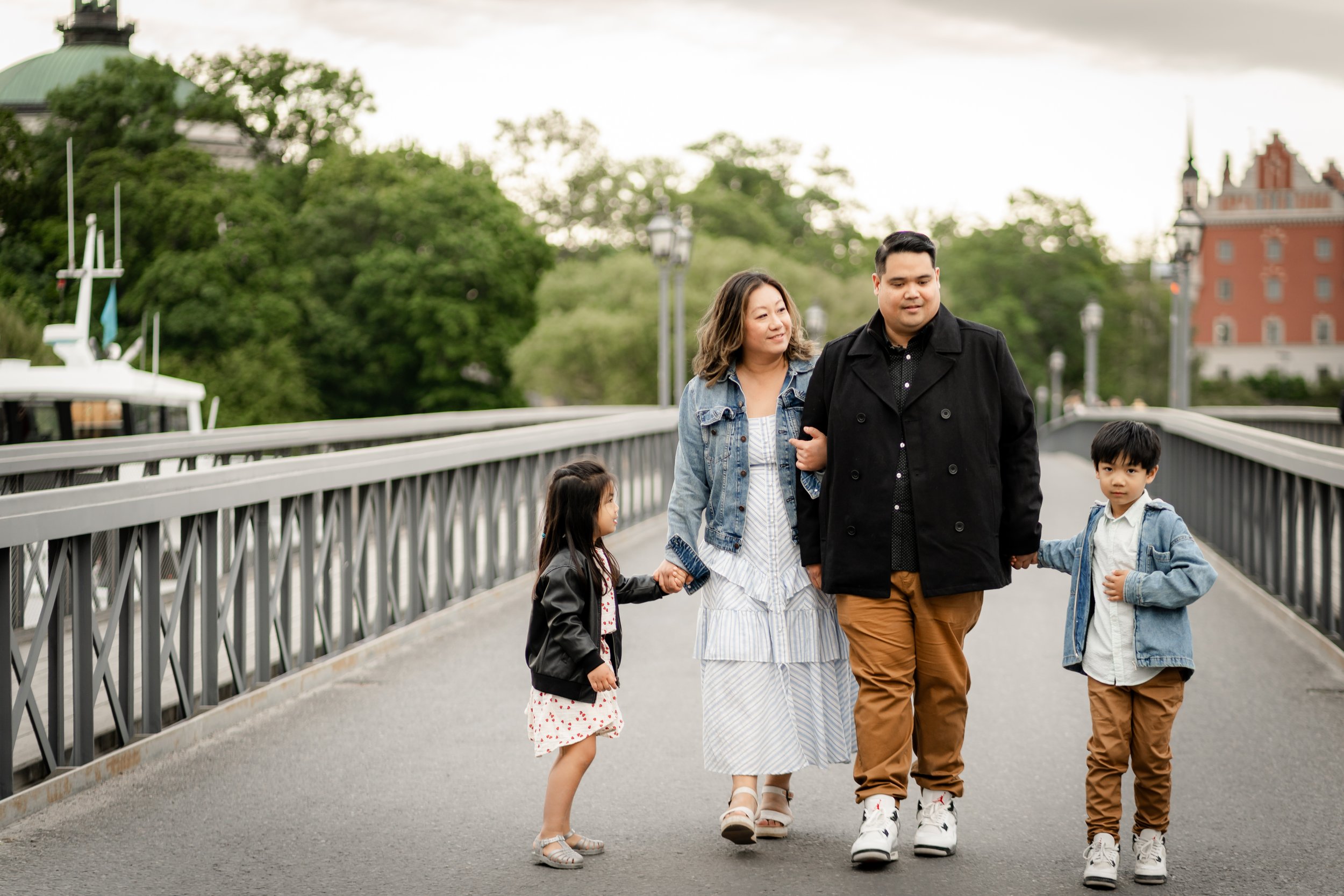 A family of four walking on a bridge, holding hands, with green trees and historic buildings in the background.