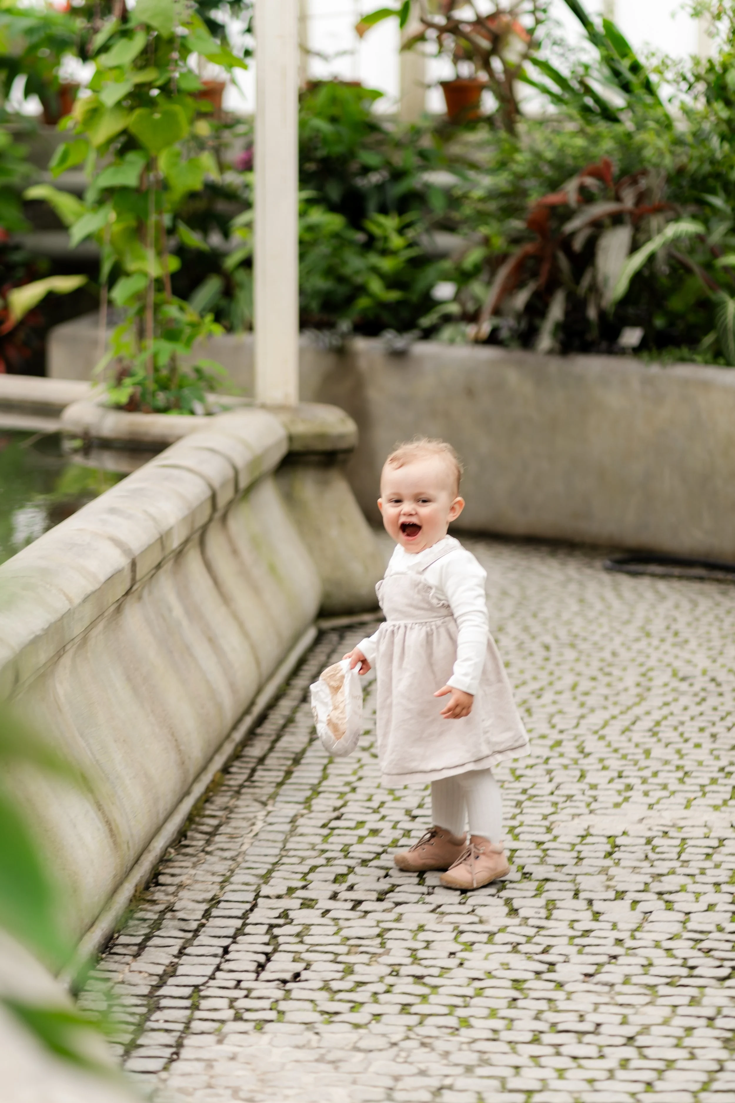 A young child with light-colored hair wearing a white dress, white leggings, and pink shoes, standing on a cobblestone path in a greenhouse or botanical garden, smiling and holding a small bag.