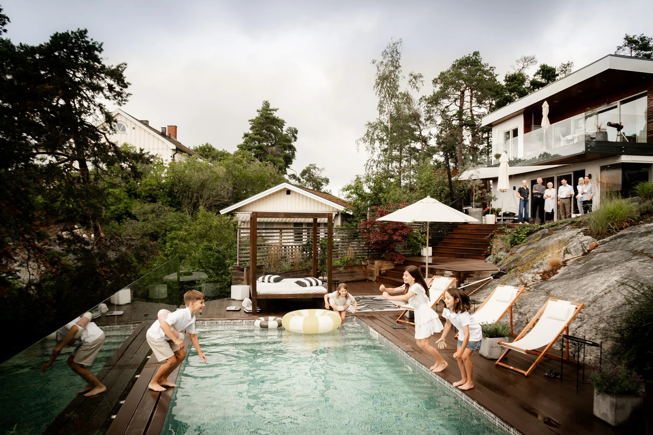 Children playing by a swimming pool with adults on the patio area above, surrounded by trees and modern houses.