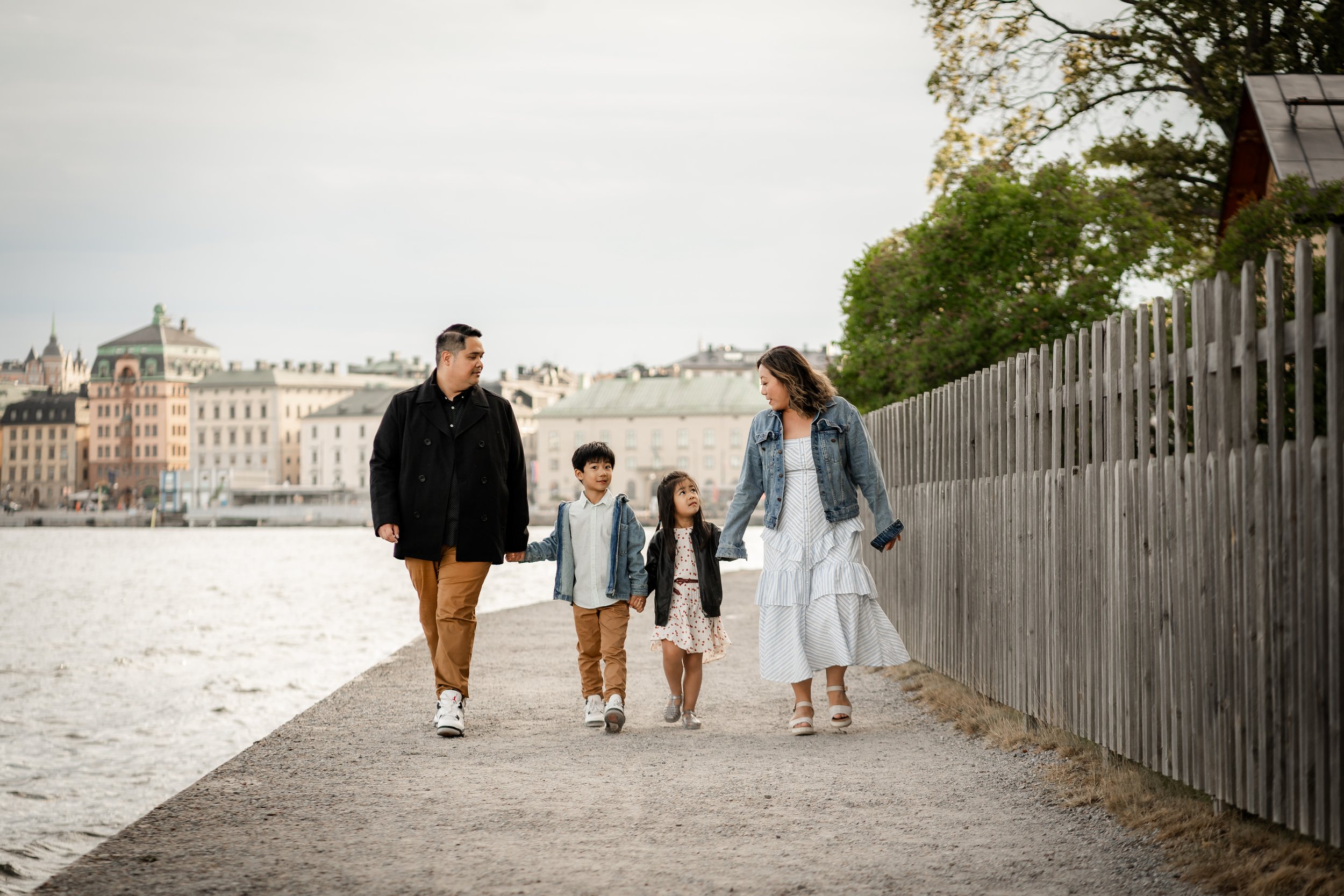 A family of four walking along a waterfront promenade, holding hands, with city buildings and trees in the background.