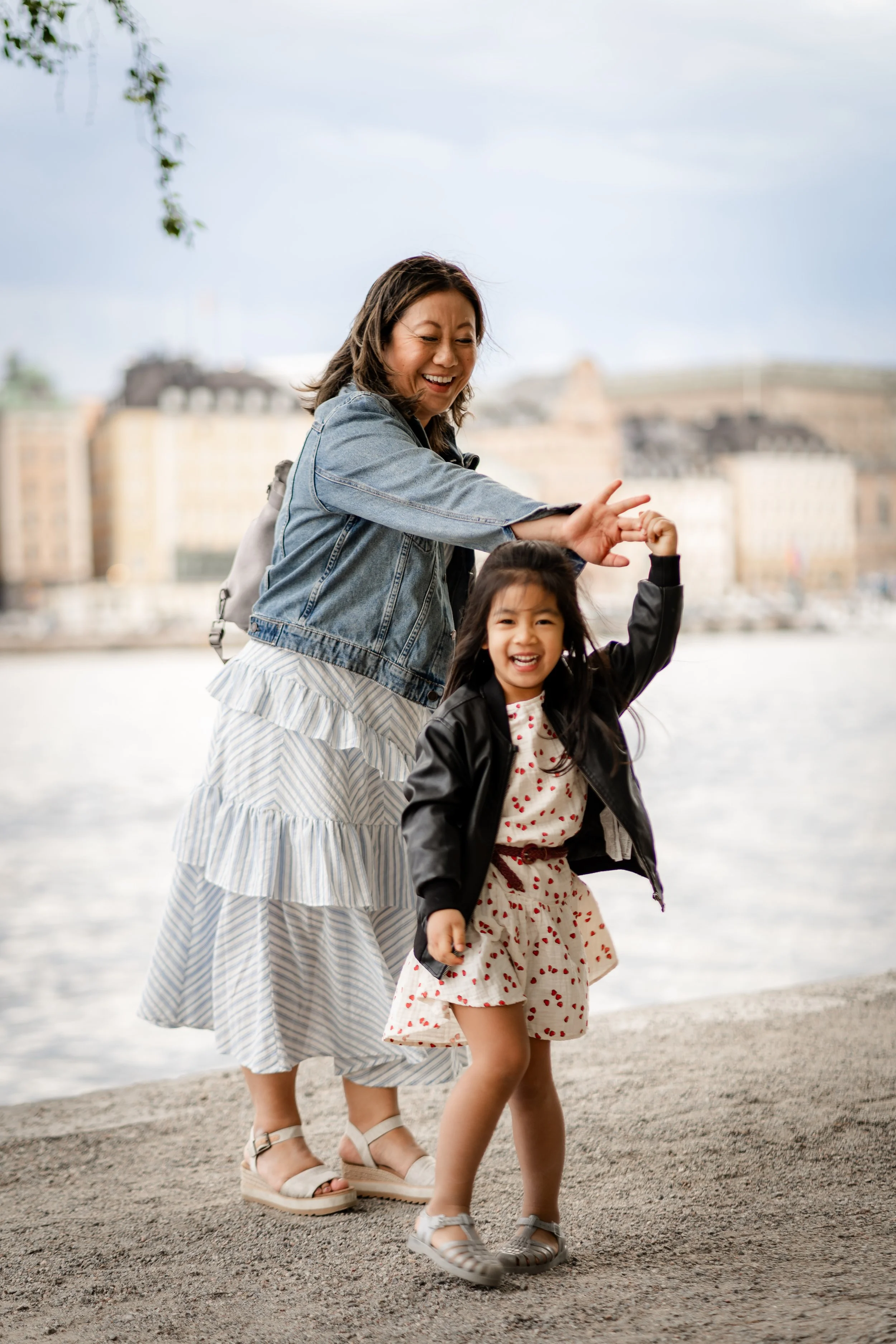 Mom and daughter smiling and playing near a waterfront, with the mom appearing to give a high five or gesture to the girl, as city buildings are blurred in the background.