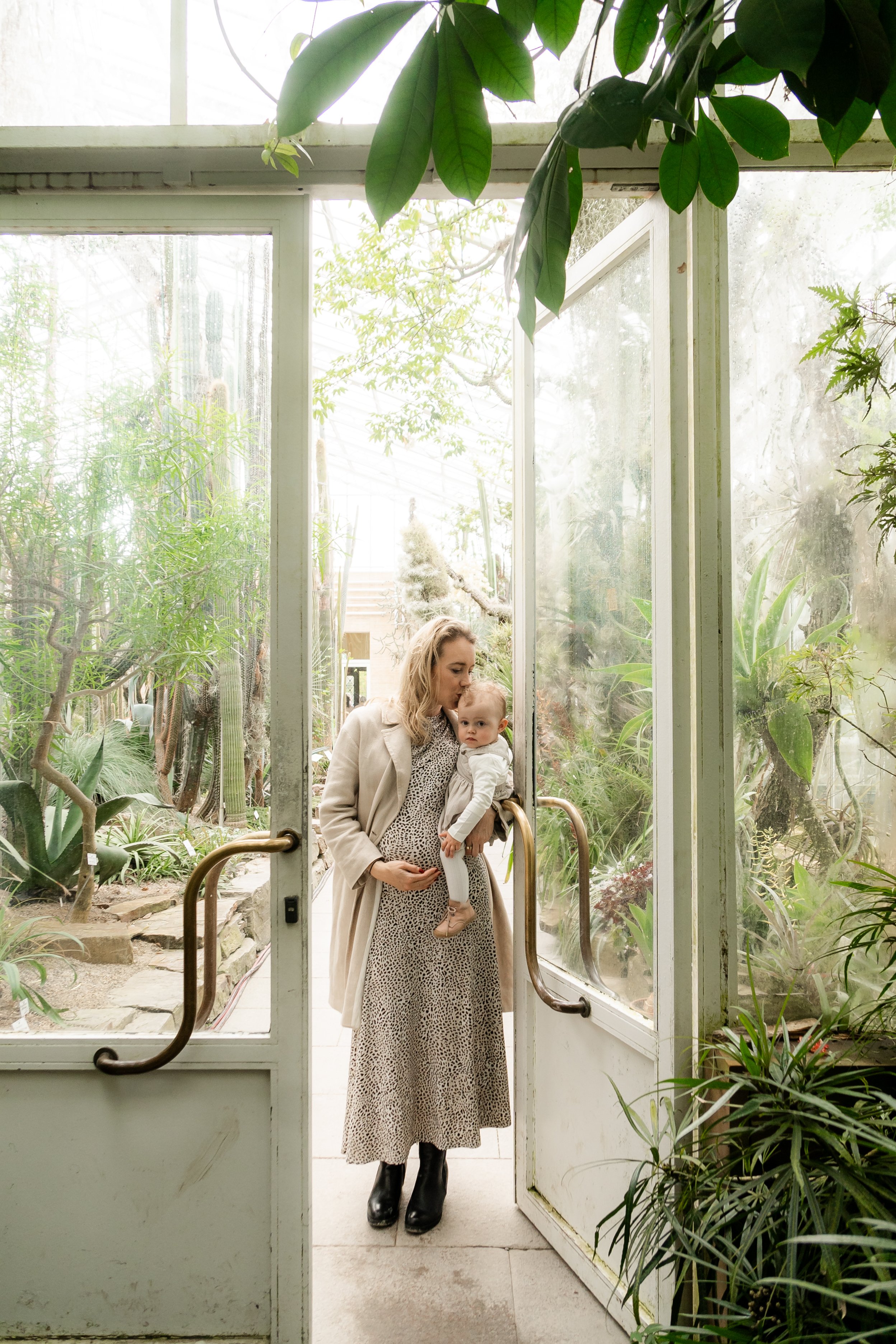A woman holding a young child at the entrance of a glass greenhouse with lush green plants inside.