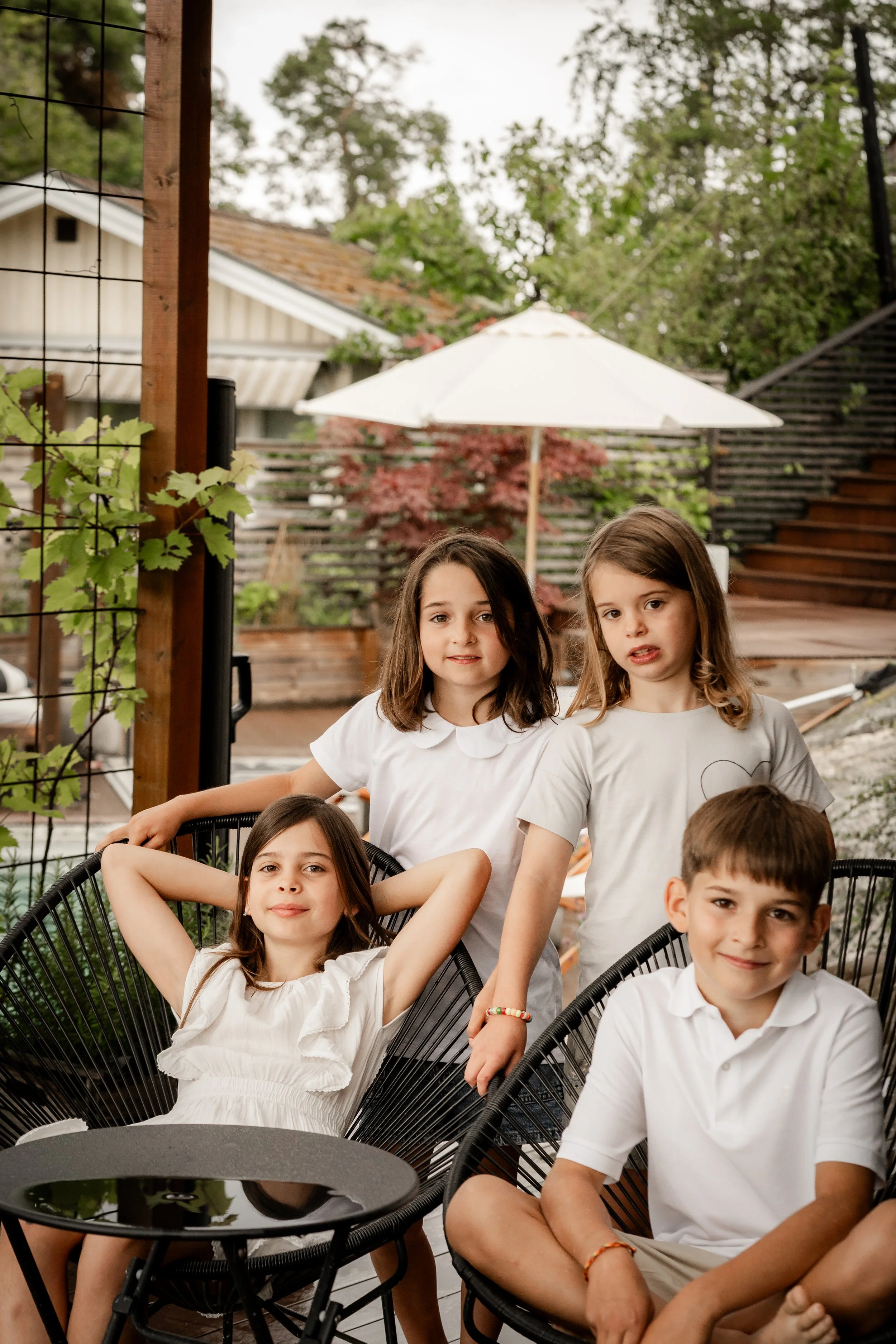 Four children relaxing outdoors in a backyard sitting and standing around black patio chairs, with a small round table, greenery, and a white patio umbrella in the background.