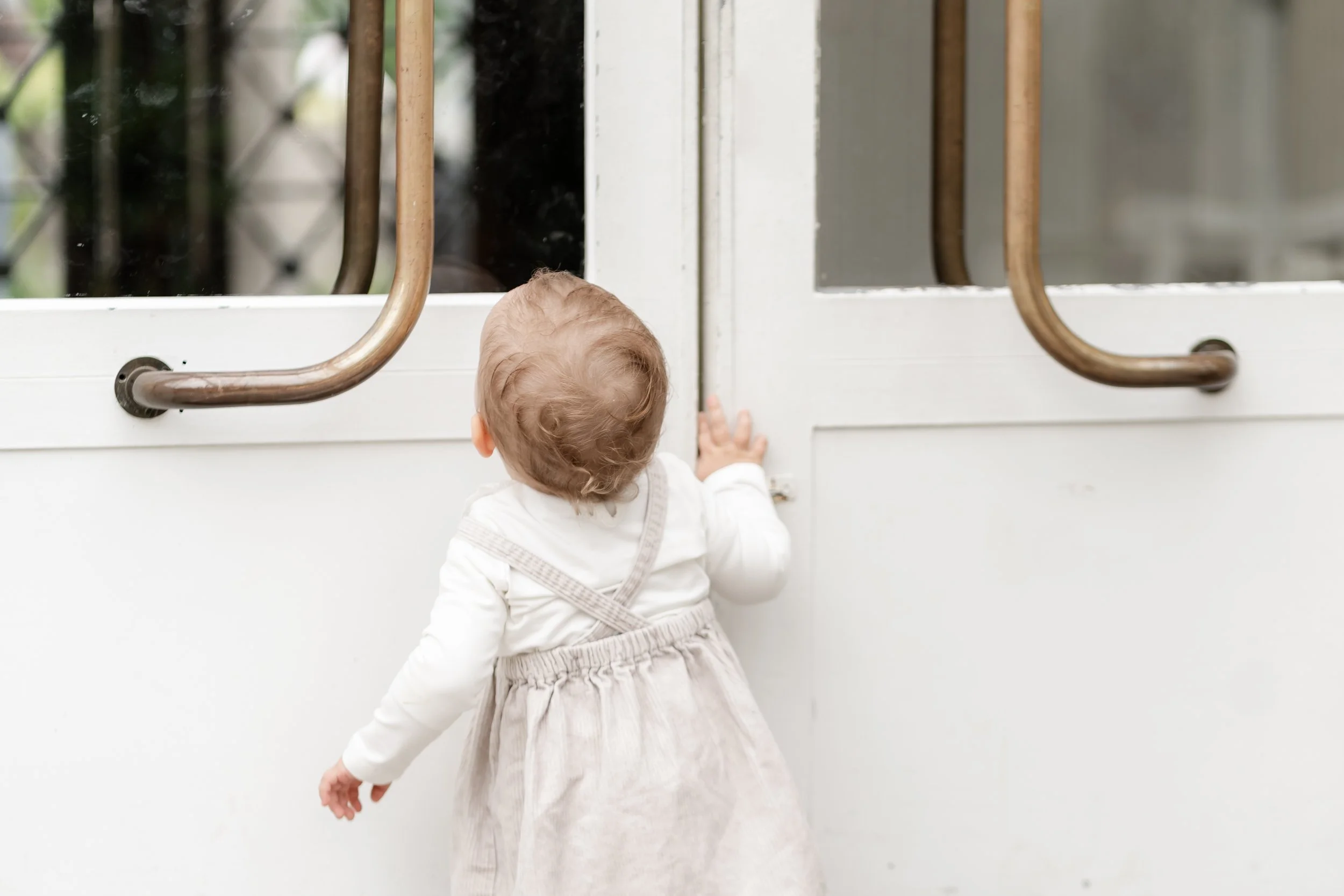 A young child with light brown hair, wearing a light-colored dress and white shirt, reaching up to open a glass door with brown handles.