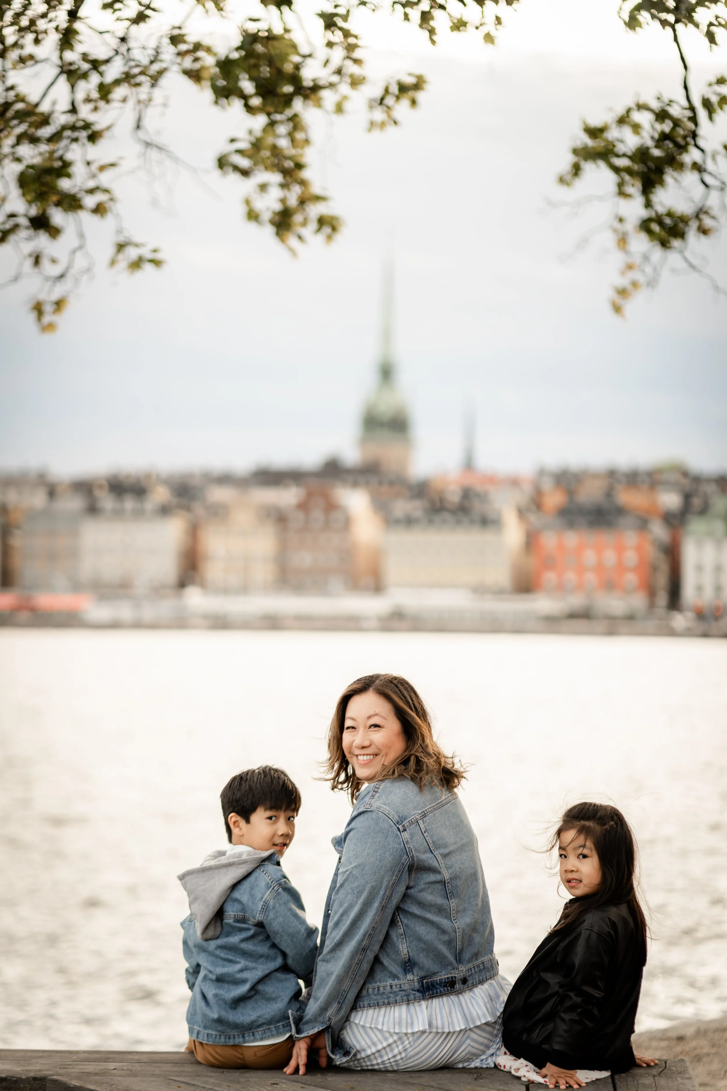 A woman and two children sitting on a dock by a river, with city buildings and a church steeple in the background.