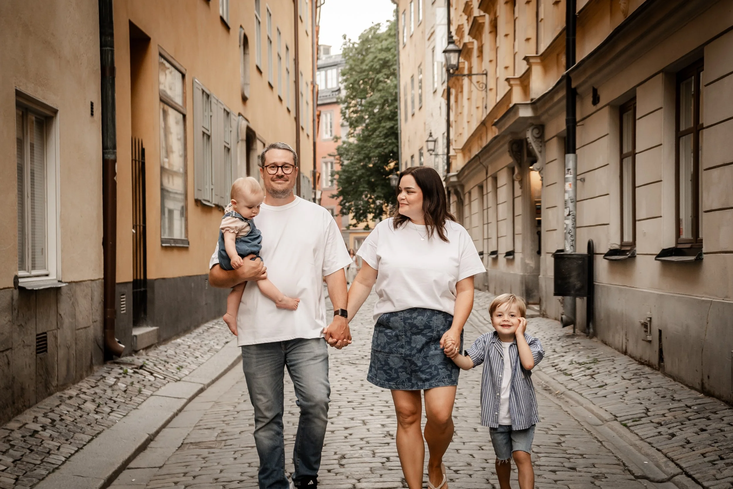 A family of four walking hand-in-hand down a cobblestone street in an urban area, with older buildings on either side.